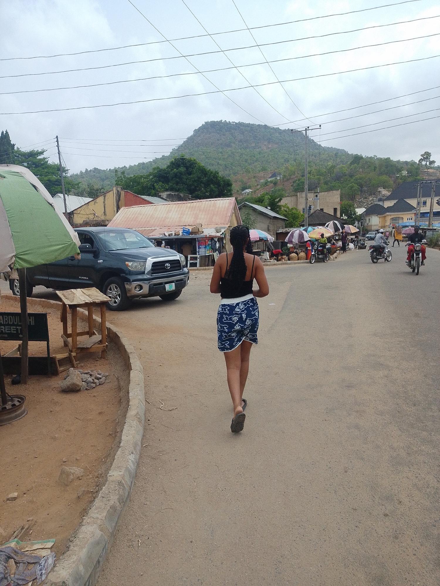 A street scene in an unidentified city shows a woman walking away from the camera on an asphalt road. She is wearing a black tank top and a blue and white patterned skirt. Several motorcycles with riders are further down the road. Vendors stand under umbrellas by the roadside. A black truck is parked on the side of the road near a wooden table. A mountain is visible in the background under a cloudy sky.