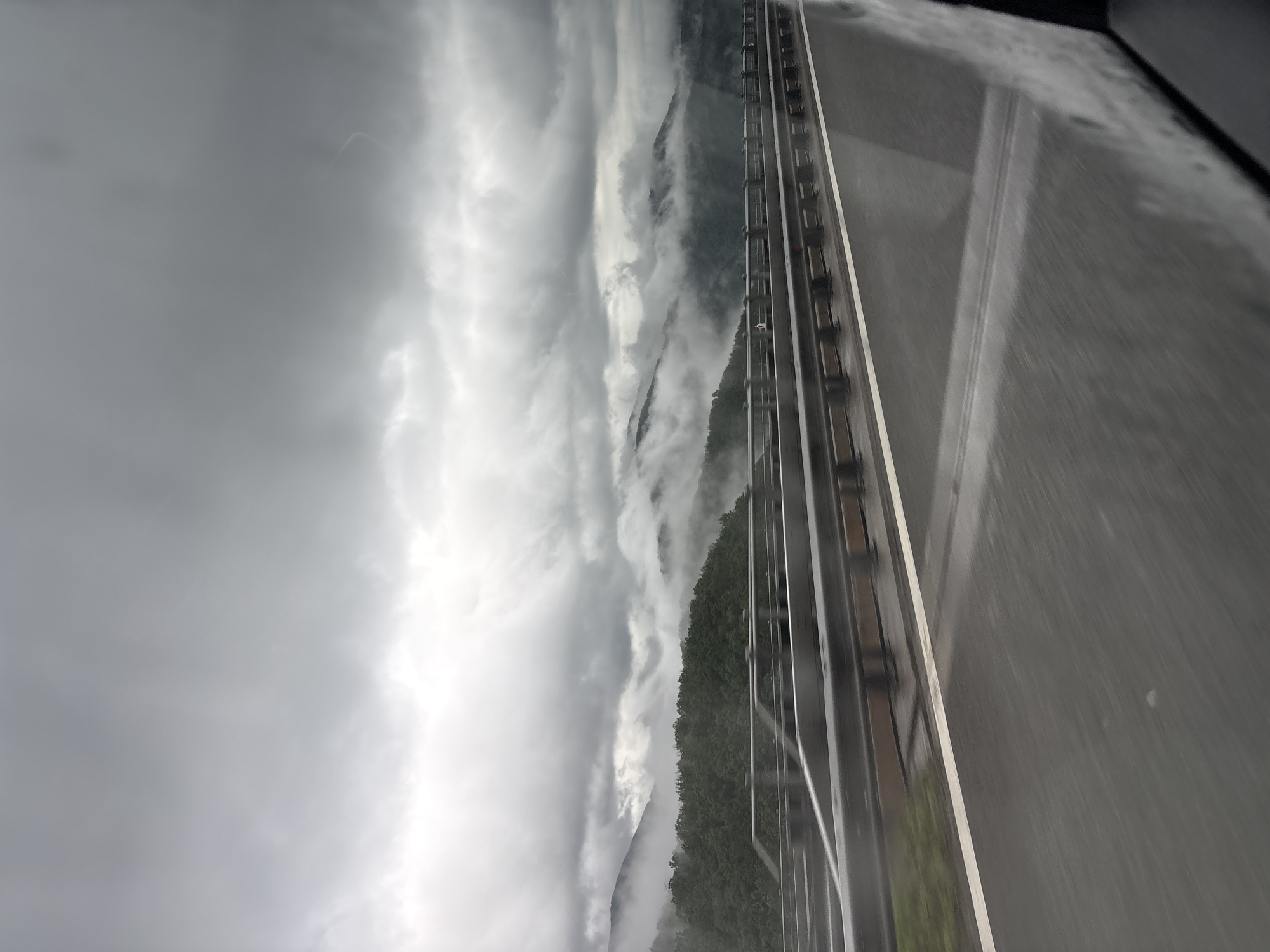 The image shows a view from a vehicle moving on a road in a mountainous region, likely in or near Vrachesh, Bulgaria. The sky is overcast with heavy clouds. Mountains, partially obscured by fog, are visible on the left side of the image. To the right of the mountains, there is a structure running parallel to the road, possibly a bridge or elevated roadway. The road surface appears wet, reflecting the dull light. The interior of the vehicle is visible at the very right edge, indicating the picture was taken from inside the car.