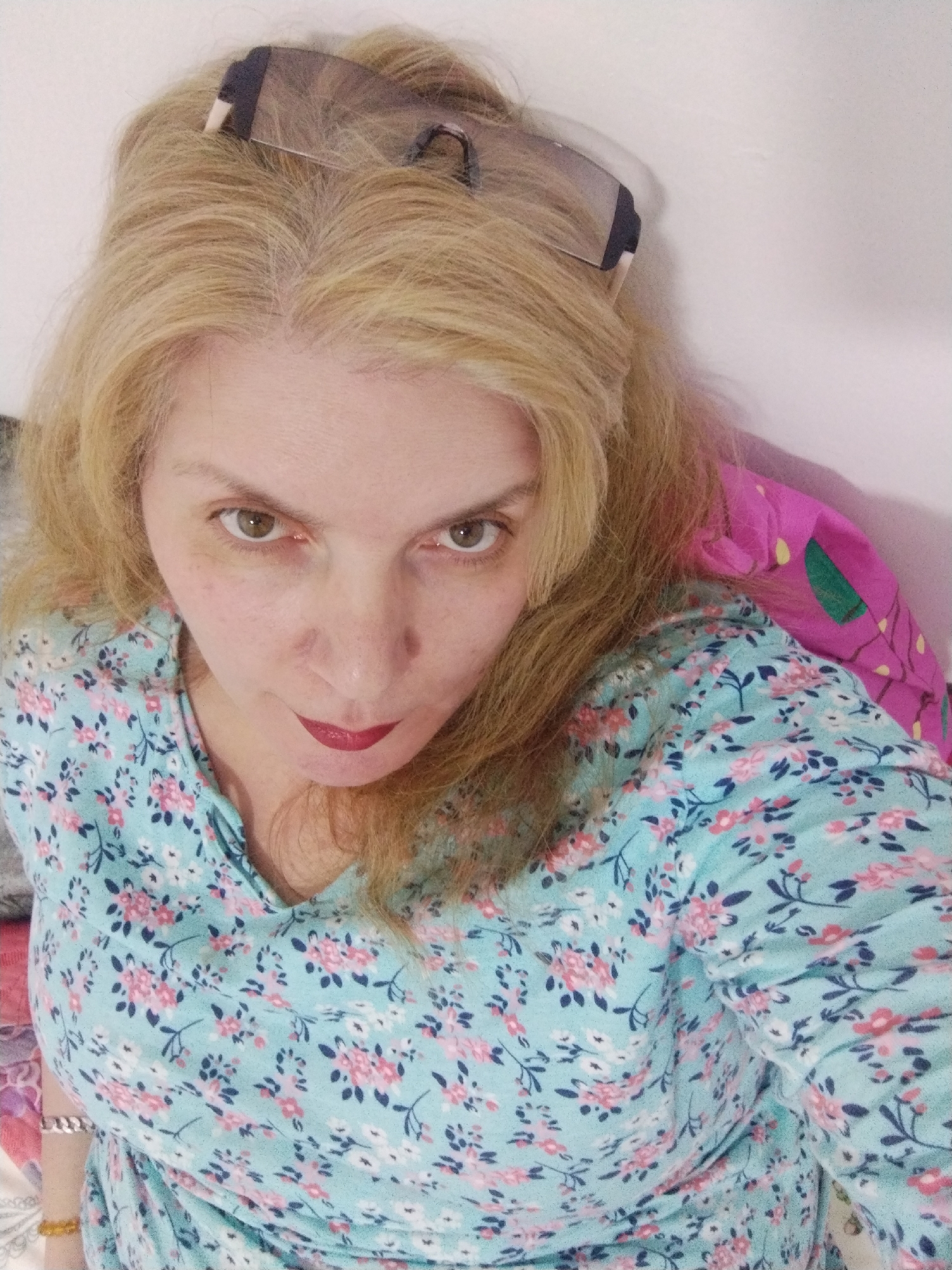 A high-angle, medium shot captures a fair-skinned woman with blonde hair, wearing sunglasses perched on her head, and a light blue floral-patterned shirt. She is positioned against a plain, off-white background, with a brightly colored pink and green patterned textile visible behind her right shoulder. The woman's gaze is directed forward, with red lipstick accentuating her lips. The lighting is diffused, casting soft shadows. No specific location in Al Mas'oudi, United Arab Emirates, can be determined from the image.