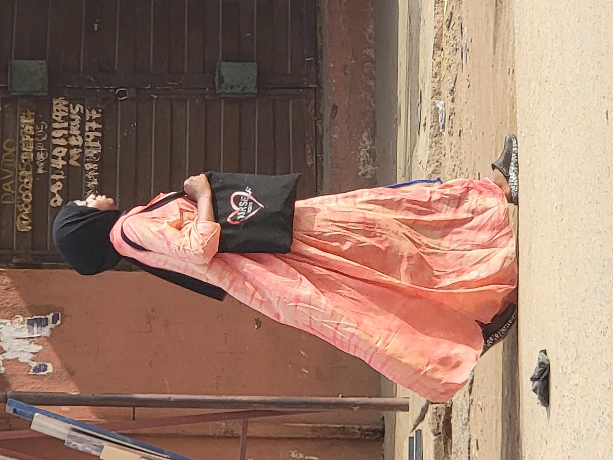 A woman wearing a black hijab and a long, patterned pink and yellow dress is walking on a dirt-covered street next to a building. She is carrying a black tote bag with a heart and the word "YOURSELF" printed on it. The building has a large brown metal door with graffiti written on it. The location is Maiburiji, Nigeria.