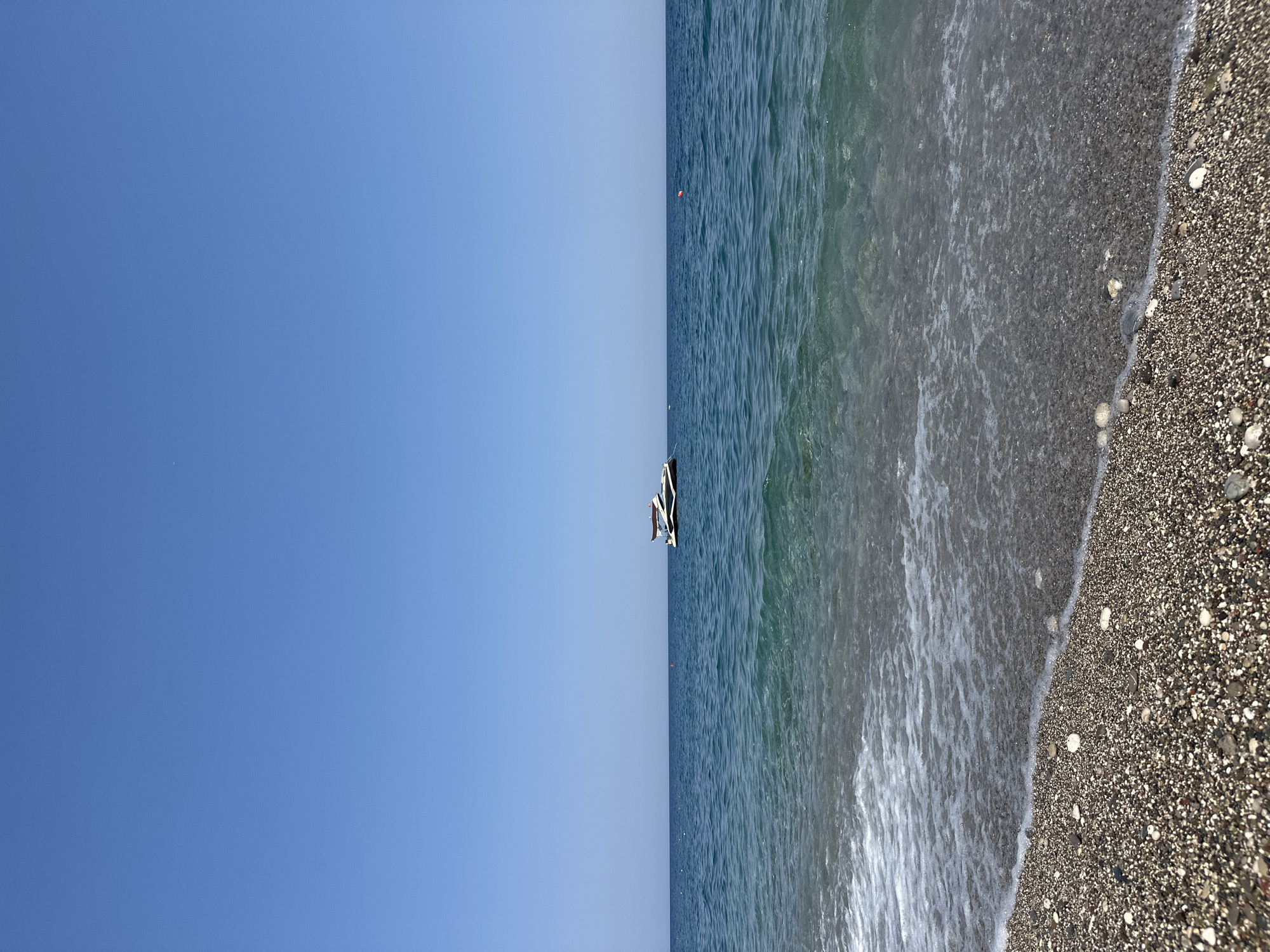 A bright, clear, sunny day at a pebble beach in Kemer, Turkey. The sea is a vibrant blue-green, with gentle waves lapping at the shore. The beach is covered in smooth, colorful pebbles. In the distance, a sleek, modern white yacht with a brown canopy is anchored in the calm water. The sky is a cloudless, deep blue.