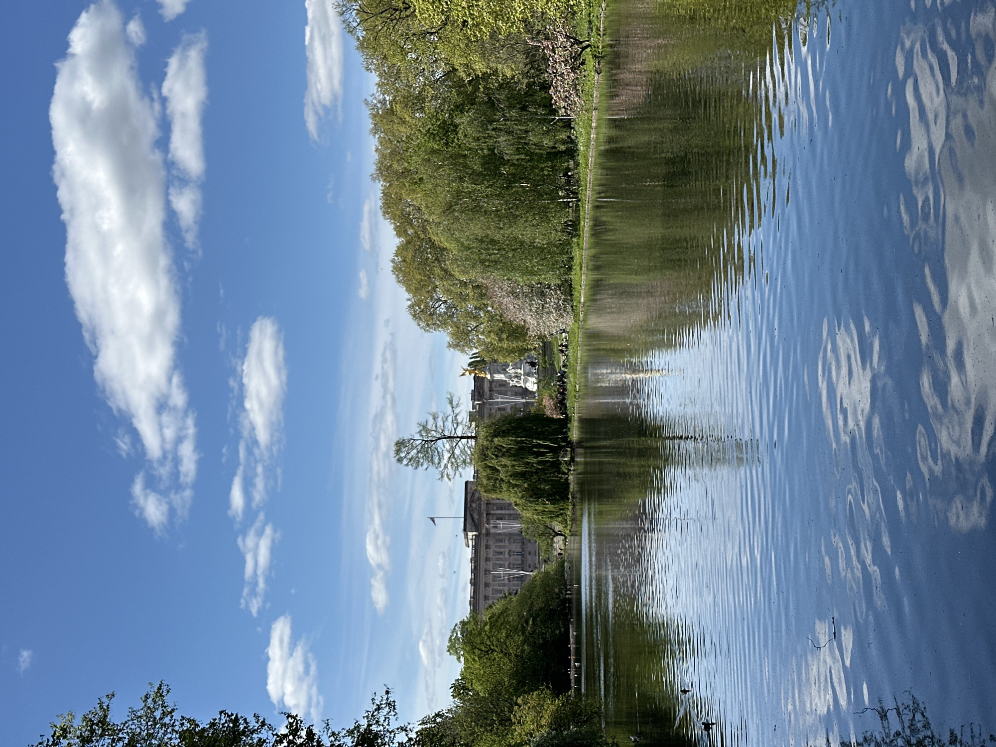 The image shows a park scene in London, United Kingdom, likely St. James's Park. In the distance, Buckingham Palace is visible, with its facade and the Victoria Memorial. A body of water, likely a lake or pond, fills the foreground, reflecting the sky, clouds, trees, and parts of the palace. The water's surface has ripples, indicating some wind. The sky is blue with scattered white clouds. Trees line the water's edge. The image is taken from a slightly low angle. There are trees in the lower part of the image, a thin line of grass separating the trees from the body of water, and then reflections of the buildings and sky in the water. The image appears to have been rotated.