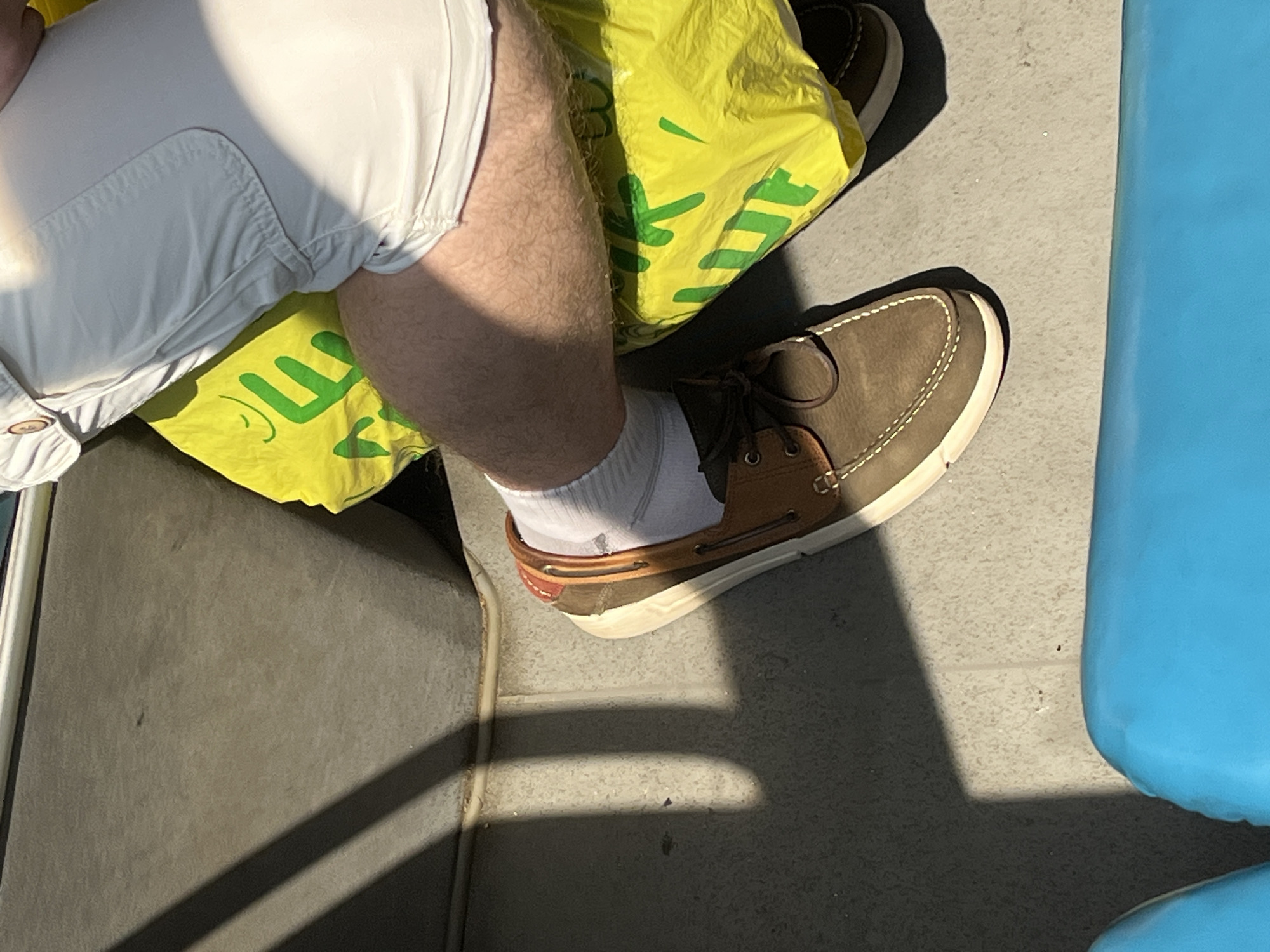 A person's legs, wearing white socks and brown boat shoes, are visible in the frame, with a bright yellow plastic bag and part of white shorts also in view. The scene appears to be indoors, possibly on public transportation, with a grey floor and seating visible. The lighting suggests it is daytime, with sunlight casting shadows. The location is identified as Minsk, Belarus.