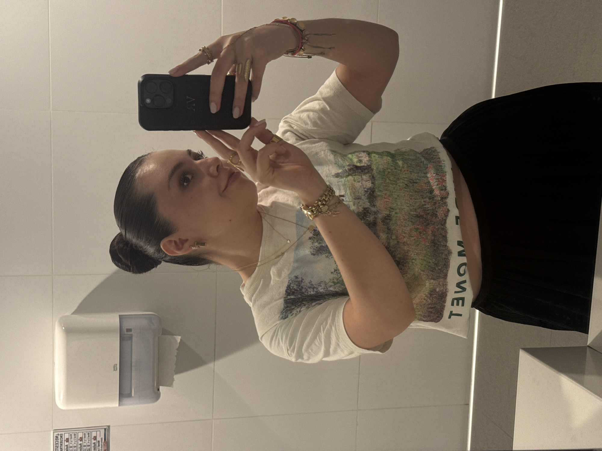A woman is taking a selfie in a bathroom with white tile walls and a large mirror. She is wearing a white t-shirt with a landscape print and black pants. She has her hair pulled back in a bun and is wearing multiple rings, bracelets, and a necklace. She is holding a black phone in her right hand and posing with her left hand. A white paper towel dispenser is visible on the wall behind her. The location is likely in Mexico City, Mexico, but there is no additional information from the image to specify a particular location.