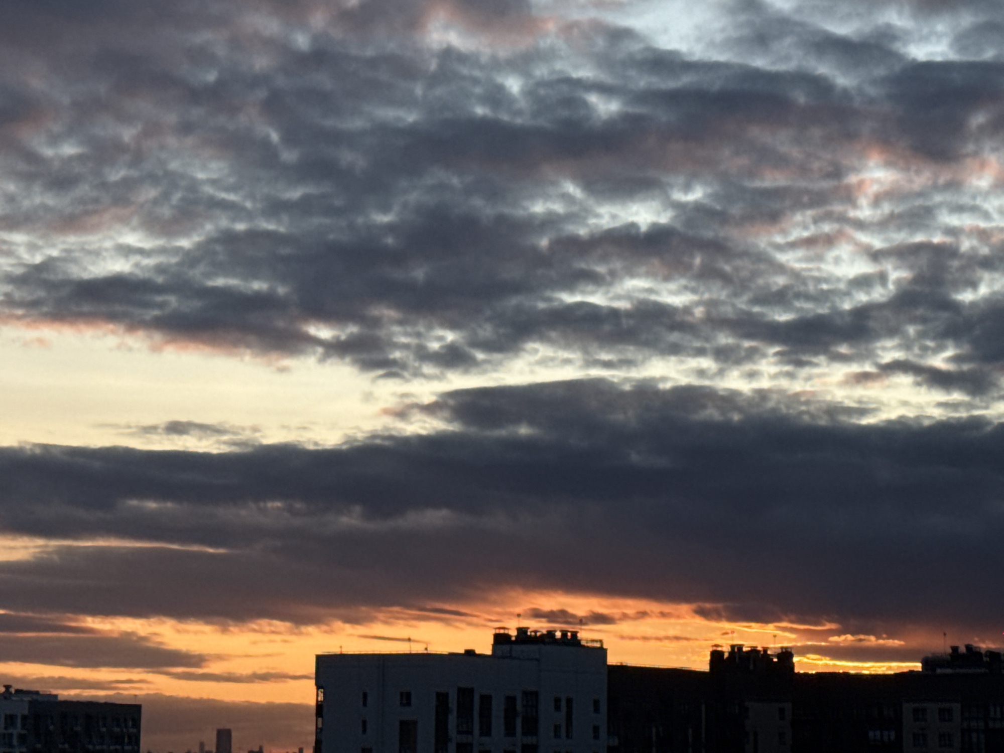 The image shows a sunset over the city of Lyubertsy, Russia. The sky is filled with dramatic clouds, with shades of gray, pink, and orange. The lower part of the image features the silhouettes of buildings, including a prominent white building with dark windows. The overall scene is one of urban twilight, with the last rays of sunlight illuminating the clouds. There are no people or animals visible in the image, and no specific actions are depicted.