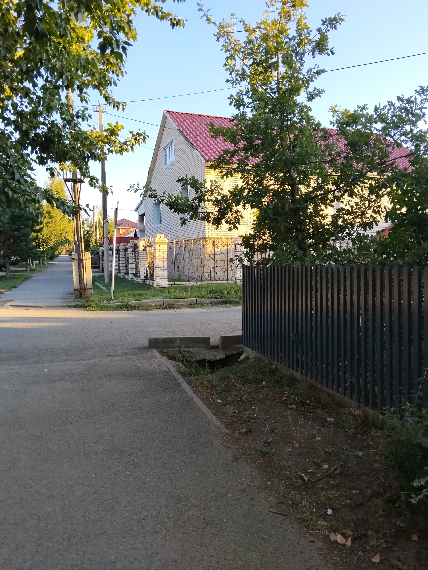 The image shows a street scene in Kokshetau, Kazakhstan. A paved road and sidewalk run along the left side of the image. On the right side, there is a dark metal fence, a patch of dirt and grass, and a brick and stone fence with an ornate metal gate. Behind the fences, a two-story house with a red roof and light-colored brick siding is visible. Trees with green leaves are scattered throughout the scene, with sunlight filtering through them. The sky is clear and blue. There are no people visible in the image.