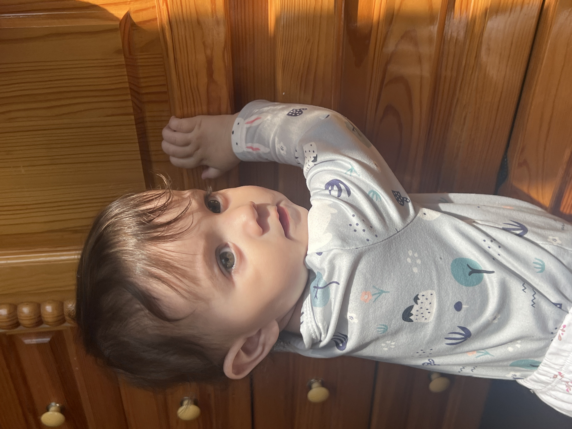 A young child, estimated to be between 12 and 24 months old, with dark brown hair and light-colored eyes, is depicted standing indoors. The child wears a light blue, long-sleeved garment patterned with stylized white, blue, and orange natural elements, and white lower attire. The child's left hand grips the vertical edge of a light brown wooden panel, possibly a door frame or wardrobe, while the right hand rests on a horizontal wooden surface, likely a drawer or cabinet top. To the child's left, a medium brown wooden dresser or cabinet with at least two visible light-colored, circular knobs is present. A strong, warm light source, consistent with direct sunlight, illuminates the scene from the right, creating distinct highlights and shadows on the wooden surfaces. The child's head is turned to the right, and the gaze is directed off-frame. The scene is situated within a residence in the city of Elena, Bulgaria.