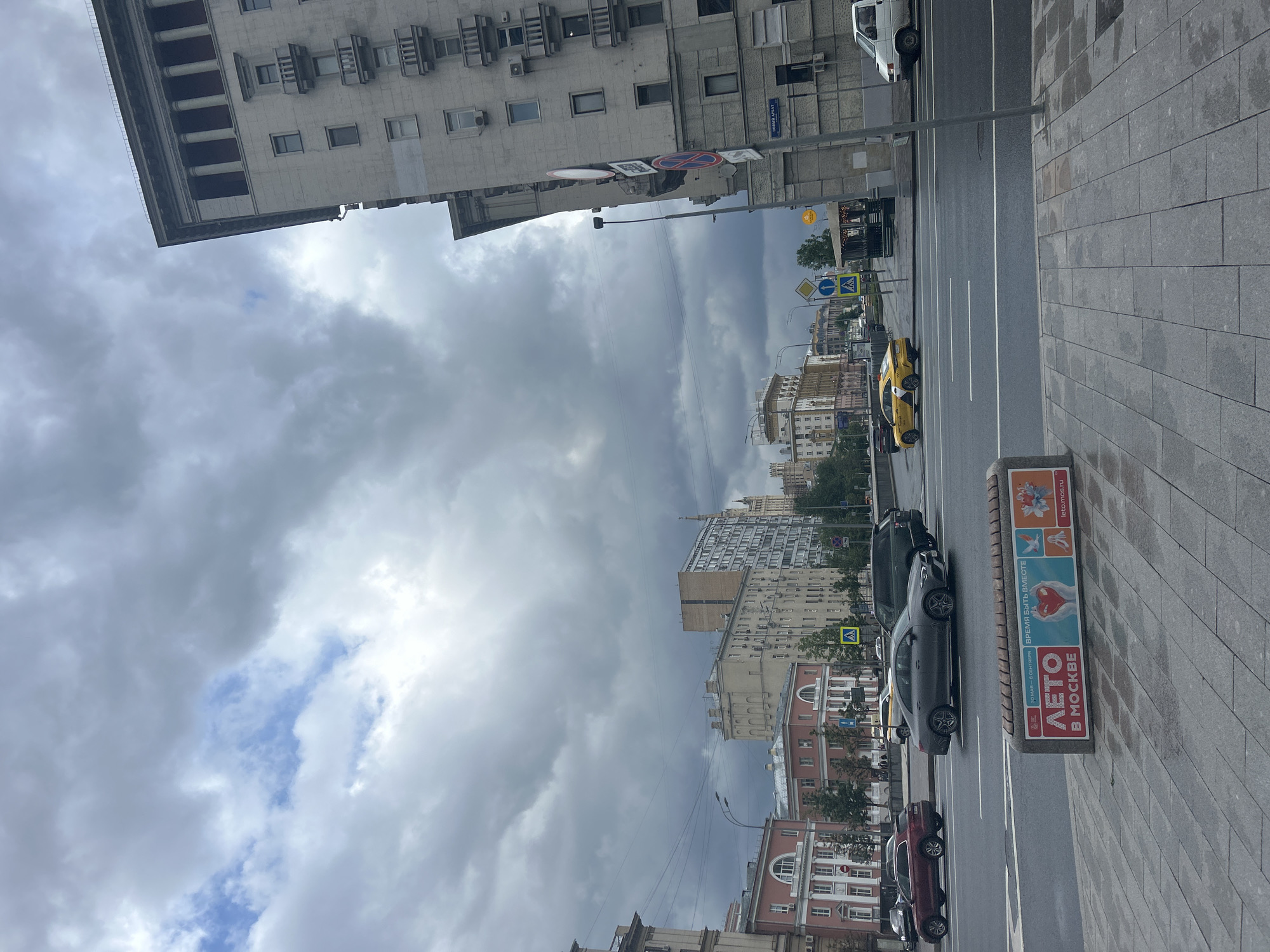 The image shows a street scene in Moscow, Russia. The photo is taken from a pedestrian perspective on a sidewalk paved with grey rectangular stones. A street runs parallel to the sidewalk, with several lanes of traffic. Cars are visible on the street, including a yellow taxi, a dark grey sedan, and a dark red car. A white van is parked on the opposite side of the street. In the foreground, a rectangular advertising panel displays a series of images and text, including the phrase "Лето в Москве" (Summer in Moscow). Tall buildings line both sides of the street, with varying architectural styles and heights. The sky is overcast with dark, heavy clouds, though there are small patches of blue visible. Street signs and crosswalks are present, indicating pedestrian crossings. The overall impression is of an urban environment with damp or recently wet conditions, as the pavement appears slightly shiny.