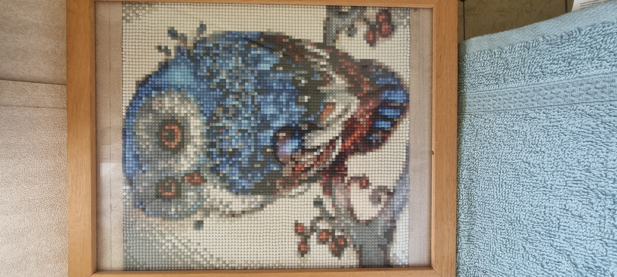 A framed diamond painting of an owl is displayed on a textured wall. The owl is rendered in shades of blue, gray, white, and red, with its eyes a striking orange. The painting is enclosed in a simple wooden frame. Below the framed artwork, a light blue textured towel is visible, partially obscuring the bottom of the frame and the wall. The background wall has a light, speckled texture. The scene does not provide any specific information about the city of Naaldwijk, Netherlands.