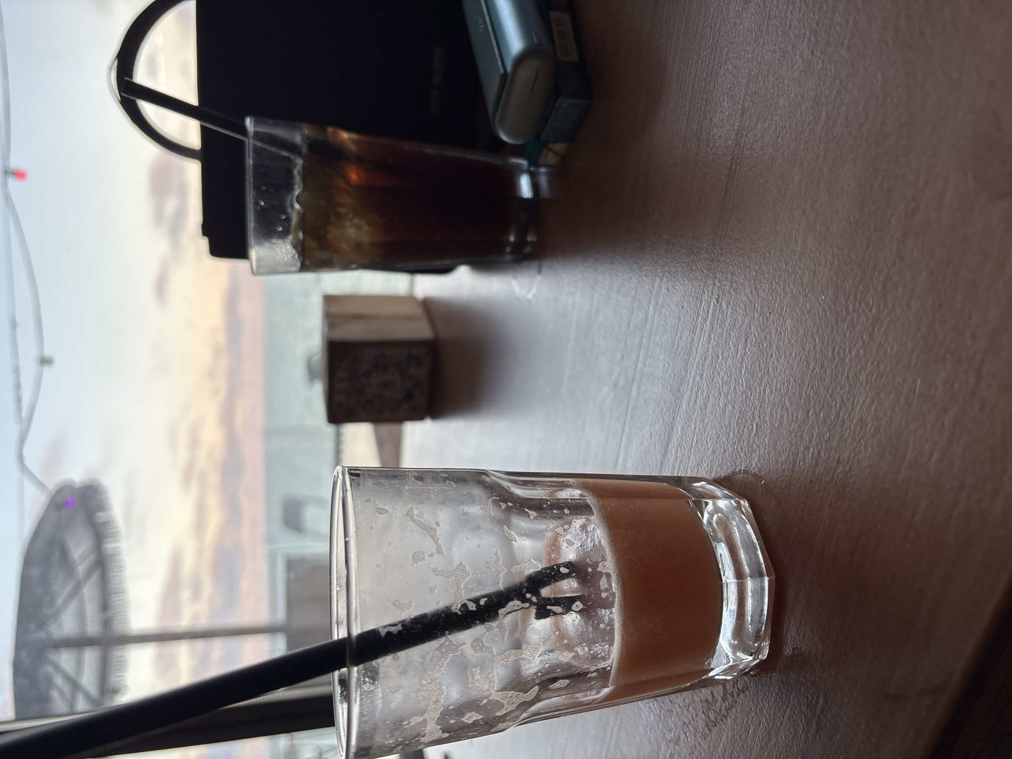 The image shows two used glasses on a table with a view of the Varna skyline through a window. One glass contains a small amount of orange liquid and a black straw. The other glass contains a dark liquid. A black electronic device is positioned on the table near the glasses. The table surface appears to be a light brown color. The Varna skyline visible through the window includes a Ferris wheel and a sunset or sunrise.
