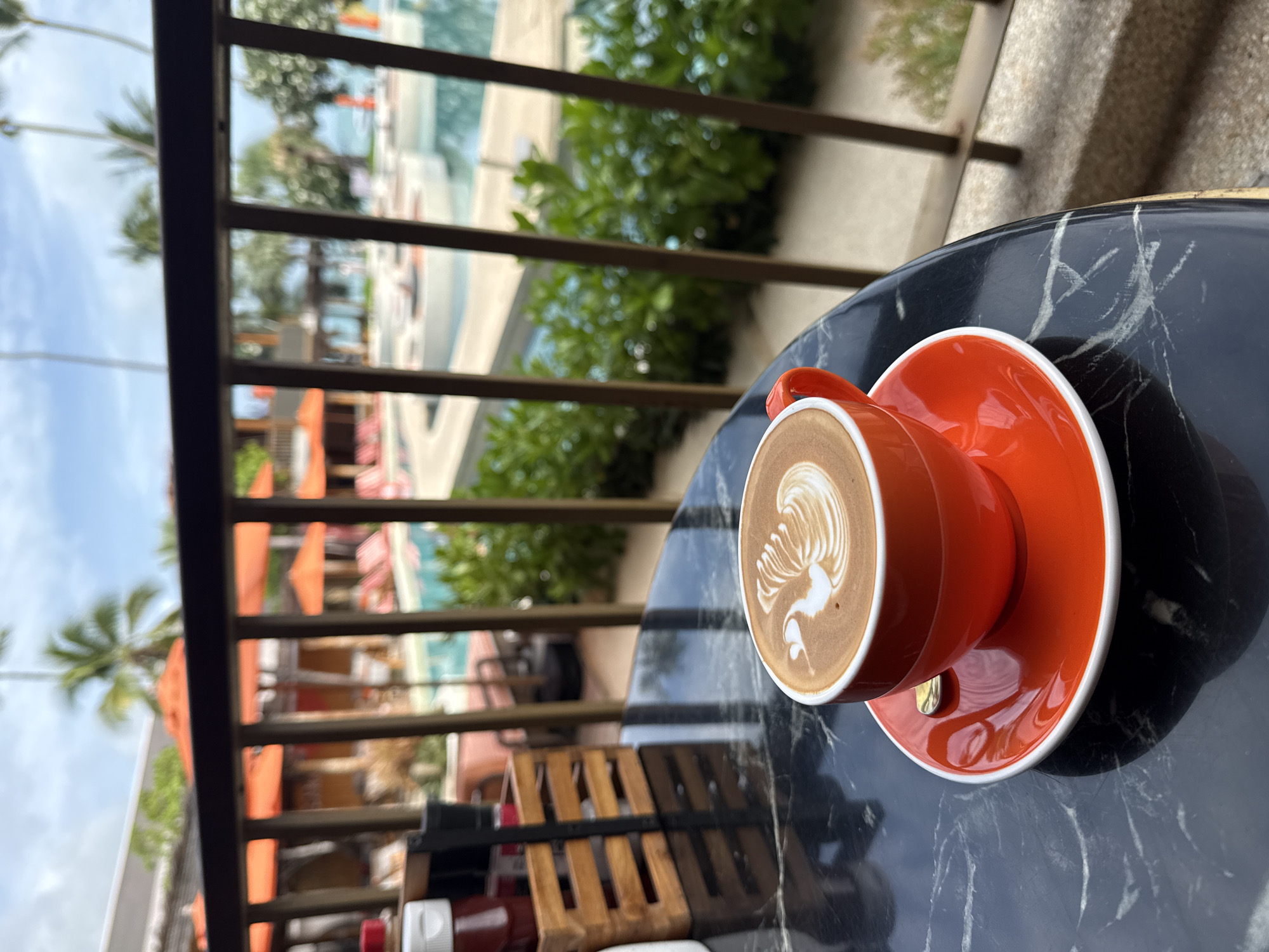 An overhead shot depicts a dark marble table with a bright orange cup of latte art coffee. The coffee is topped with white foam art, resembling a swan. Beside the cup, a small golden spoon rests on the saucer. In the background, a railing overlooks a resort-style swimming pool and the ocean. Palm trees and lounge chairs are visible, suggesting a tropical setting in Ko Samui District, Thailand. The scene is captured during daylight, with a slightly overcast sky.