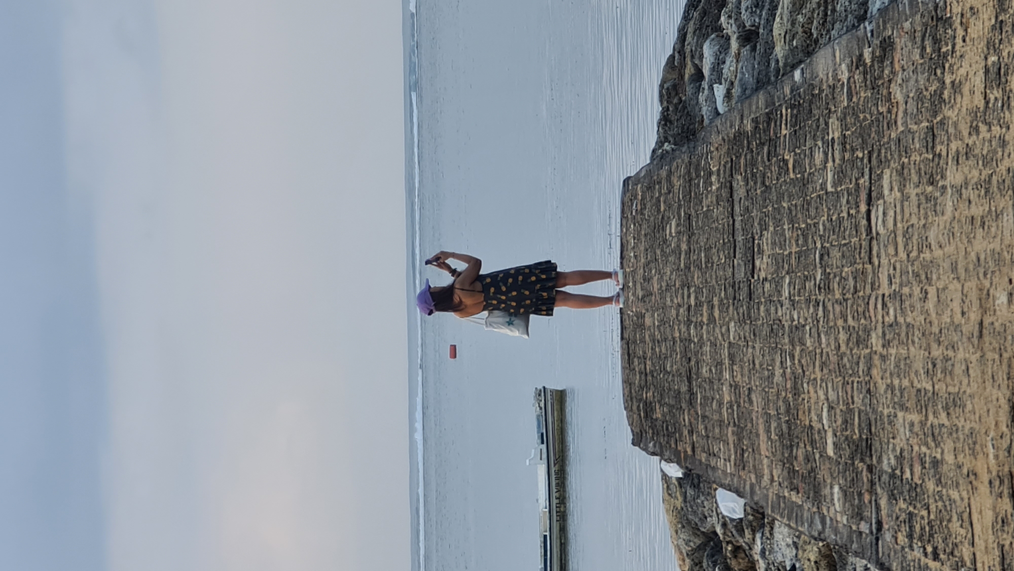 A person wearing a purple cap and a dark dress with a pineapple pattern stands on a stone pier, facing the ocean and holding a smartphone to take a picture. The person is positioned in the center of the frame, with their back to the viewer. The ocean is calm with small waves in the distance. A white boat with the words "LITTLE VIKING" visible on its side is moored to the left of the pier. An orange buoy floats in the water. The sky is overcast. The pier is constructed of interlocking stones. The image appears to be taken at the beach in Sanur, Denpasar, Indonesia.