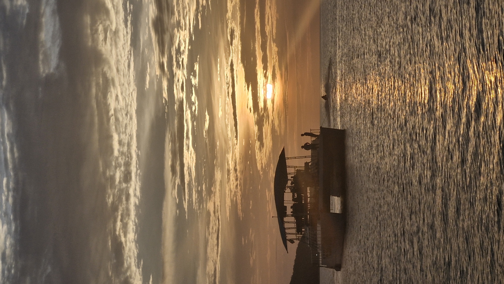 A wide shot captures a catamaran boat sailing on the ocean during sunset. The boat is silhouetted against the orange and yellow sky, its upper deck covered by a canopy. Several people are visible on the boat, standing and sitting. Two figures stand near the railing, looking out at the water. In the distance, a person on a jet ski is riding away from the boat, leaving a wake. The sun, partially obscured by clouds, casts a golden reflection on the rippling water. The scene appears tranquil, with the warm hues of the sunset creating a serene atmosphere. The location is identified as Unknown, Unknown.