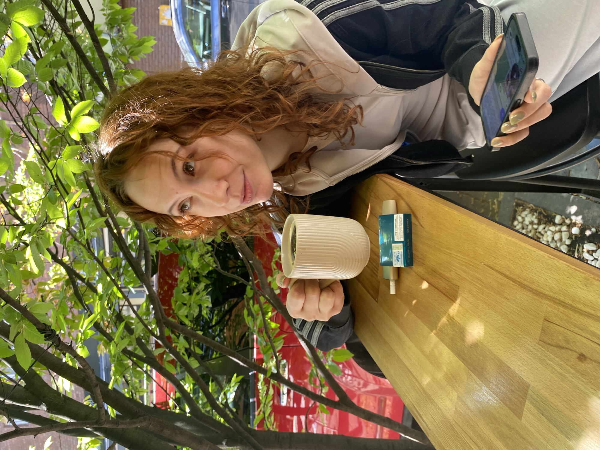 A woman with auburn, curly hair sits at a wooden table outside in Novi Sad, Serbia. She is wearing a light-colored hoodie, a black jacket, and light pants. She holds a smartphone in her right hand and a white, ridged mug in her left hand. On the table, next to the mug, is a small, turquoise-colored rectangular container. A tree with green leaves is partially visible in the foreground. The background shows glimpses of a red structure, a blue vehicle, and white pebbles. The woman is looking at the camera.