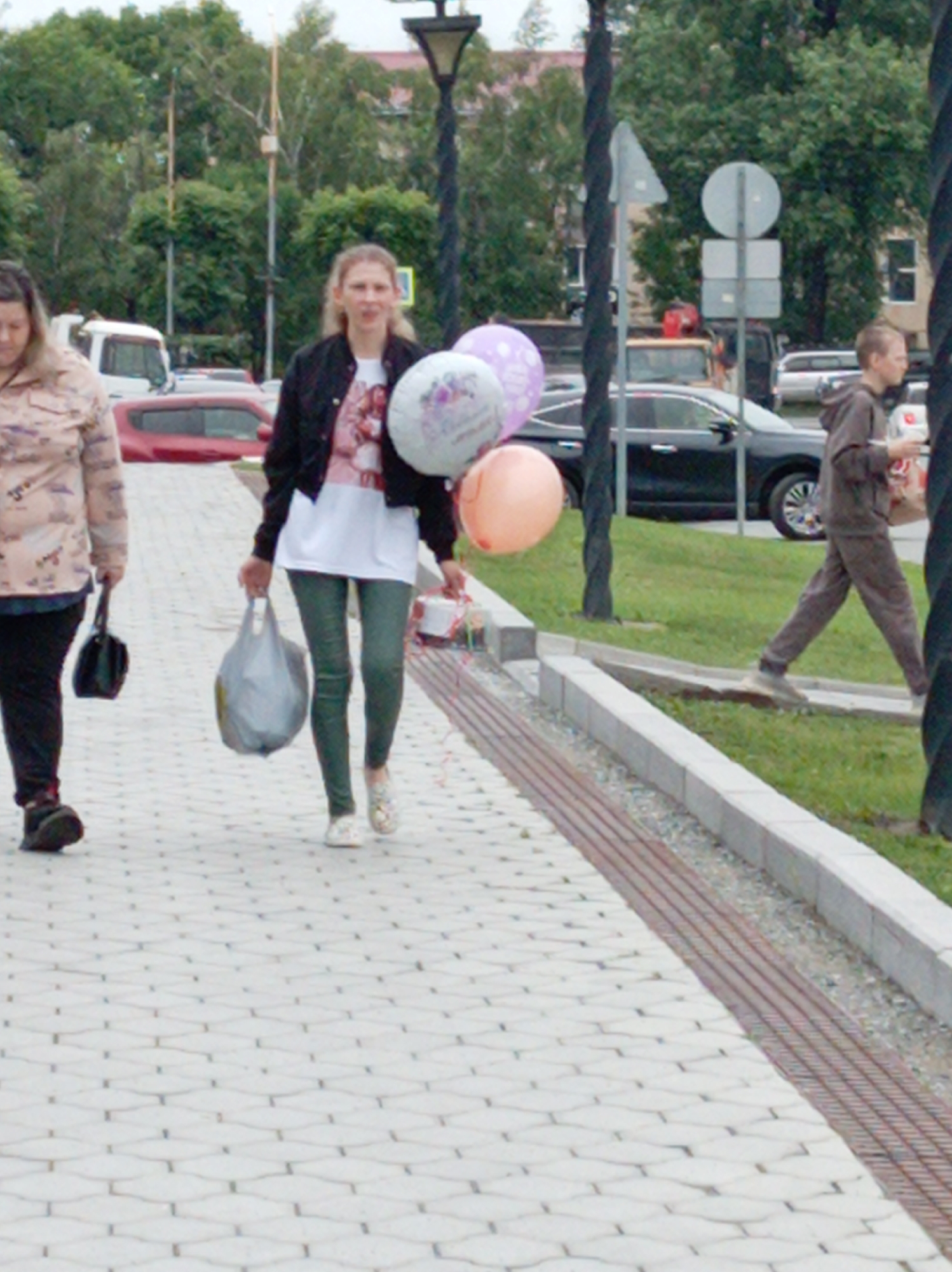In the city of Artyom, Russia, on a paved walkway, three individuals are walking. On the left, a woman with blonde hair and a patterned shirt walks with a black handbag in her left hand. To her right, a young woman with blonde hair wears a black jacket over a white t-shirt and dark green pants. She carries a large gray plastic bag in her right hand and several balloons in her left. Further to the right, a young man in a brown hooded tracksuit walks away from the camera, carrying a red and white bag. Behind the individuals, trees and buildings are visible, along with cars and street signs. The walkway is bordered by a grassy area and a drainage grate.