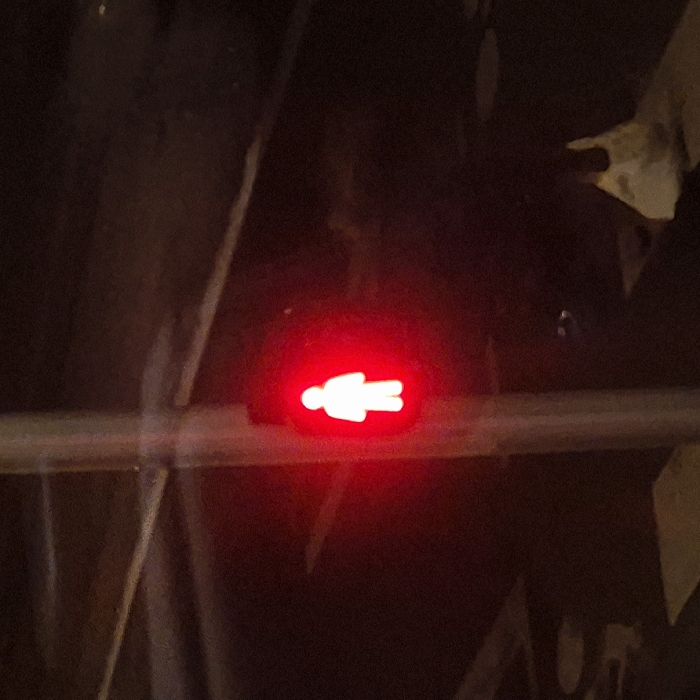A red illuminated figure of a person signifies "do not walk" at a pedestrian crossing. The light is mounted on a horizontal bar. The surroundings are dark, with indistinct structures and surfaces visible in the background. The scene is dimly lit, focusing attention on the bright red signal.