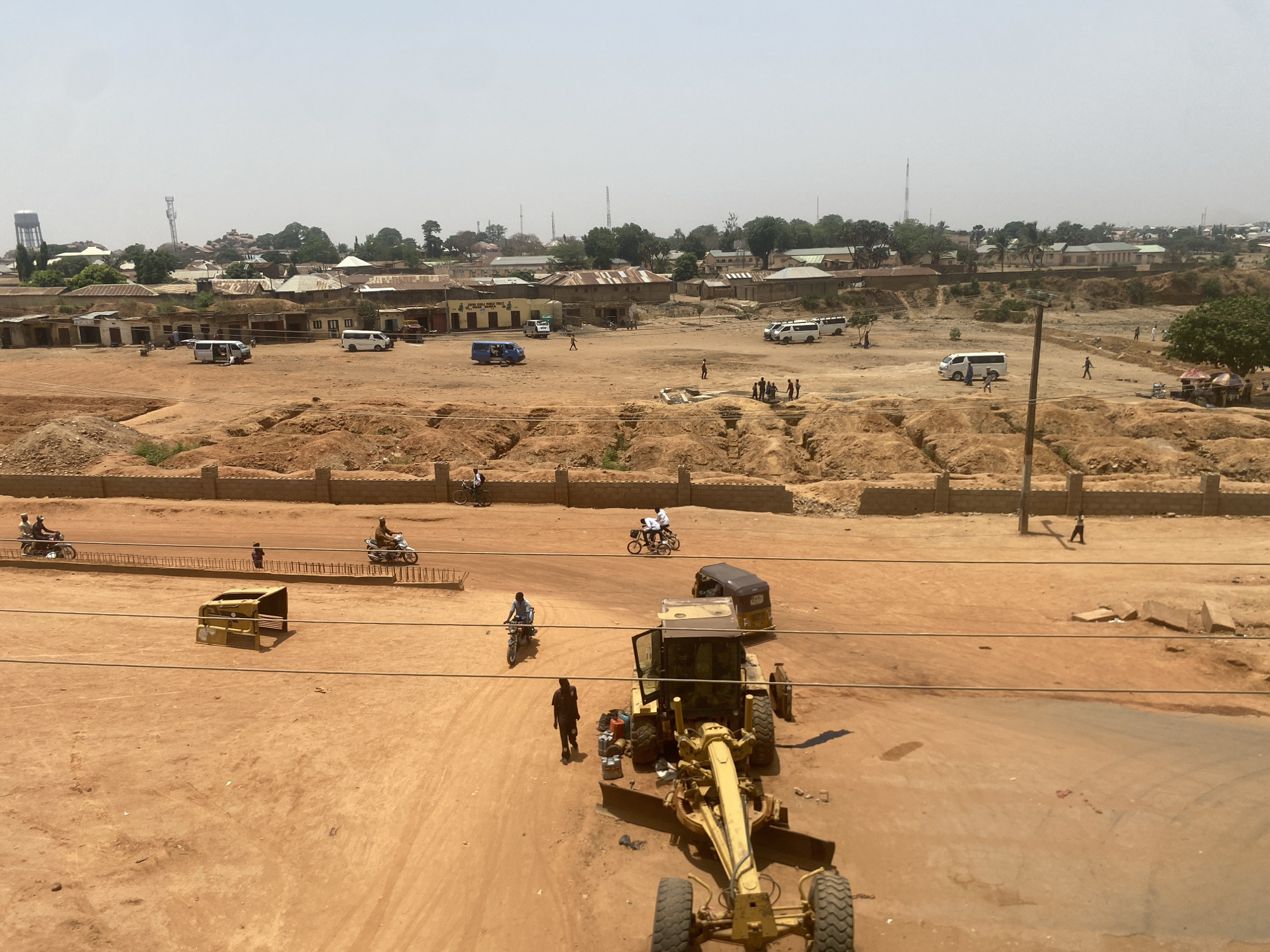 The image depicts a construction site in Tudun Wada, Nigeria. In the foreground, a yellow construction grader and other heavy equipment sit on a dirt surface. A person walks near the grader. A road runs across the middle ground, with people on motorcycles and bicycles traveling on it. In the background, there is a residential area with buildings and vehicles. A water tower is visible. The landscape is predominantly dry and dusty.