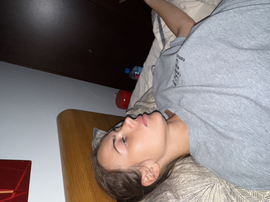 A single individual, a young adult female, is depicted lying on a bed in an indoor setting. Her head rests on a beige pillow with a light-colored leaf or feather pattern. She is seen in profile, facing towards the right side of the frame, with a neutral expression and open eyes. Her brown hair is pulled back, revealing the left side of her face and ear. She is wearing a light grey short-sleeved t-shirt with dark, faint text partially visible on the chest. A light-colored duvet or blanket covers the lower portion of her body.
The bed features a light brown wooden headboard. Behind the individual, a light grey wall is visible, with a partially visible rectangular red artwork featuring geometric lines mounted on it in the upper left corner. To the right of the individual's head, on the bed or an adjacent surface, a small, round, red object (possibly a speaker) is present. Behind this, a clear plastic water bottle with a blue cap and clear liquid is also visible. To the far right of the frame, a dark brown vertical surface, likely a wardrobe or door, is seen. The lighting in the room appears soft and diffused. No specific markers or details within the image allow for the identification of the location as Warsaw, Poland.