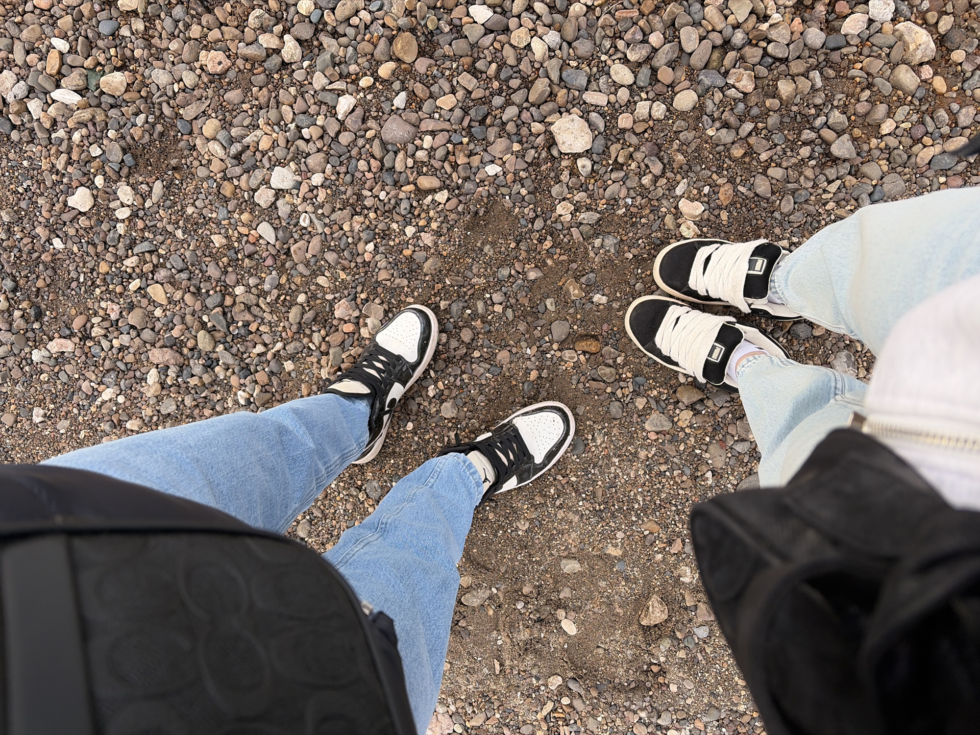 Two pairs of legs wearing jeans and sneakers stand on a surface covered in small pebbles and dirt. The pair on the left wears black and white sneakers, while the pair on the right wears black sneakers with thick white laces. Part of a black bag is visible on the left side of the image. Part of a black bag and white jacket are visible on the right. The image was taken in Chaikovsky, Russia.