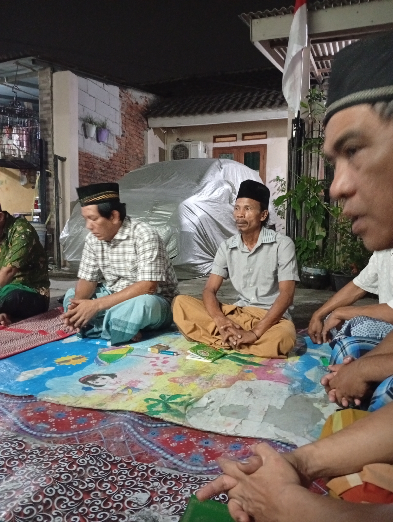 The image depicts a group of men gathered outdoors in what appears to be a residential setting in Caringin, Indonesia. The men are sitting on the ground on a colorful patterned mat. Three men are clearly visible in the foreground and mid-ground, all wearing traditional Indonesian attire including peci caps. One man in the foreground on the right is partially out of focus, with his face turned to the right. The man in the center is wearing a light-colored short-sleeved shirt and brown pants, with his hands clasped in front of him. To his left, another man is wearing a plaid shirt and light blue sarong, also with his hands clasped. In the background, a car covered with a tarp is visible, as is a wall made of brick and white plaster. A flag, possibly the Indonesian flag, is mounted on a pole to the right. The lighting suggests it is evening or nighttime. The environment appears to be a simple outdoor living area, possibly a yard or patio.