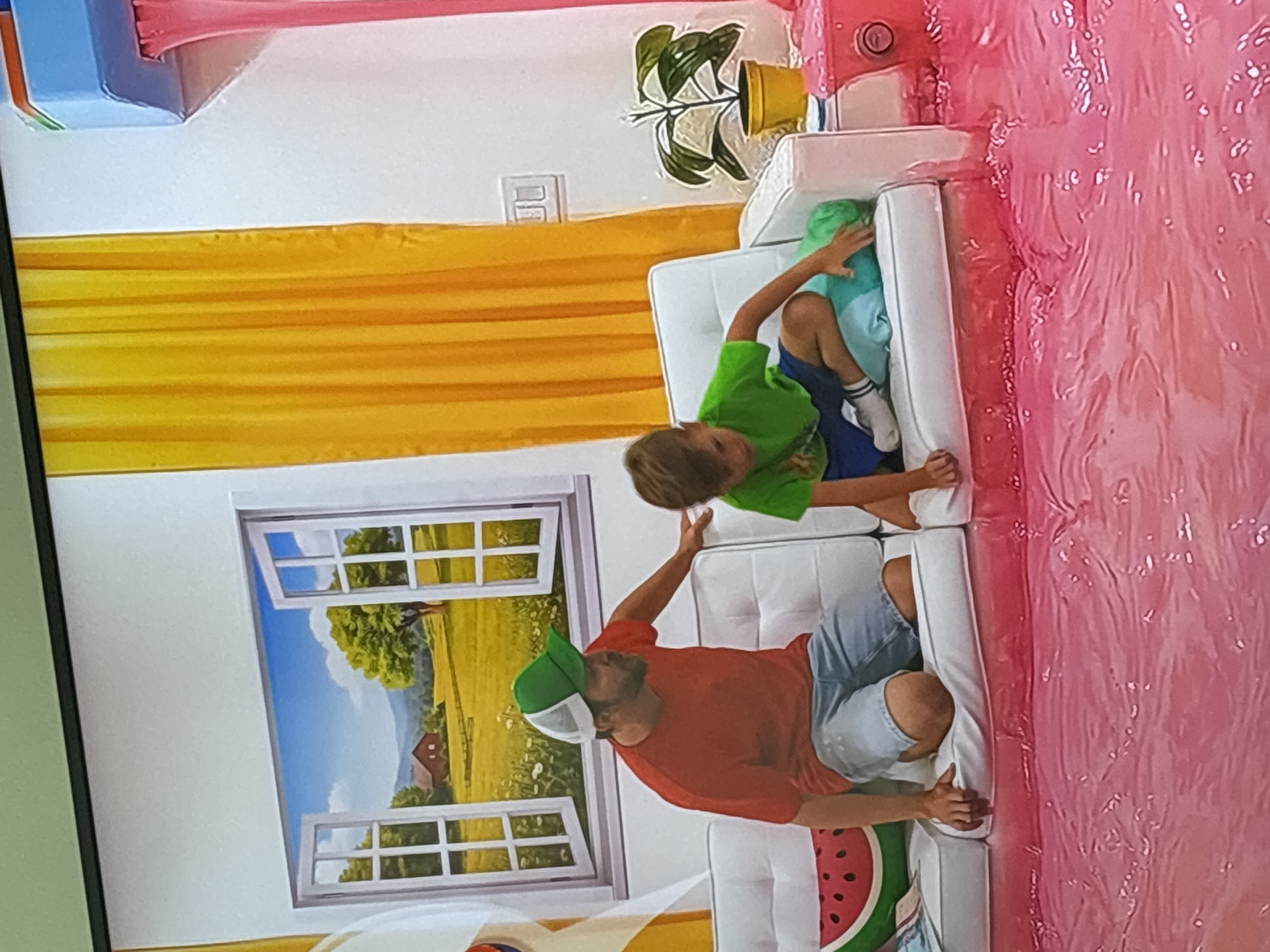 This is a high-angle, full-shot, eye-level photograph of a man and a boy on a white couch in a room. The man is wearing a red shirt, a green and white hat, and denim shorts. The boy is wearing a green shirt and blue shorts. They are sitting on the couch, which is surrounded by a pink, liquid-like substance. The background features a painted landscape with a barn and trees visible through an open window, as well as yellow curtains and a pink structure with pink liquid dripping from it. The image appears to be taken in the "Museum of Illusions" in Dubai, United Arab Emirates, a popular tourist attraction known for its interactive exhibits that create optical illusions.