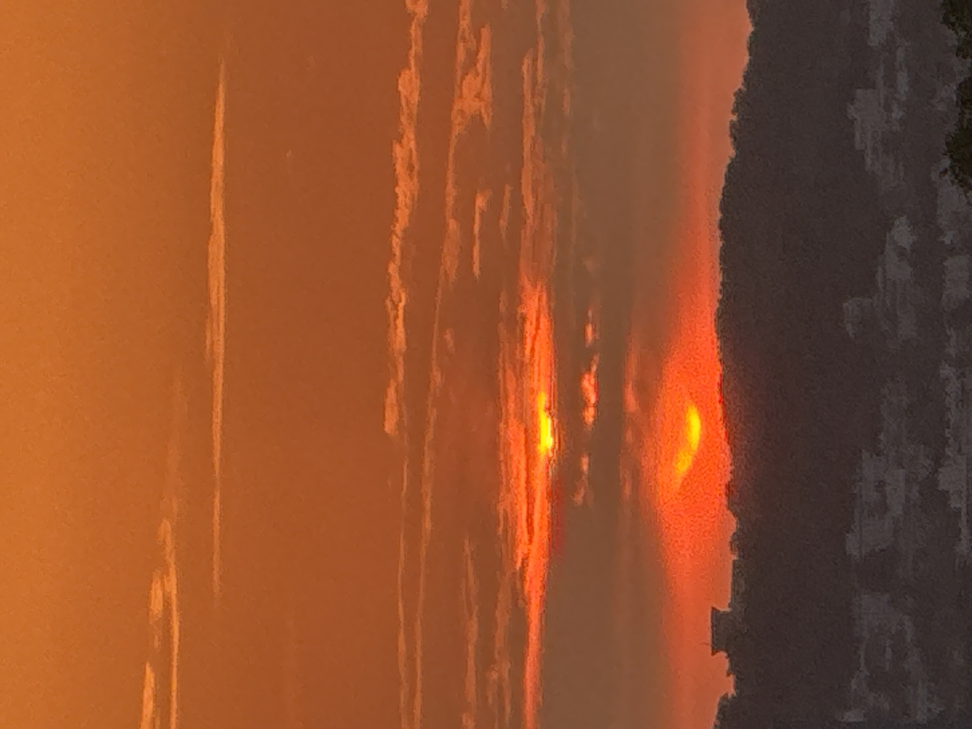 The image captures a sunset over a city. The sky is filled with warm orange and red hues, with scattered clouds. The sun, partially obscured by clouds, appears as a bright orange orb. In the foreground, a dark, silhouetted landscape suggests rolling hills or a mountain range, with the faint outlines of buildings and structures visible. One prominent building, possibly a tower or a complex, stands out against the horizon. The lower portion of the image reveals a more detailed view of city buildings and structures, rendered in muted tones against the darkening landscape. The overall impression is one of a serene and dramatic sunset unfolding over an urban environment.
