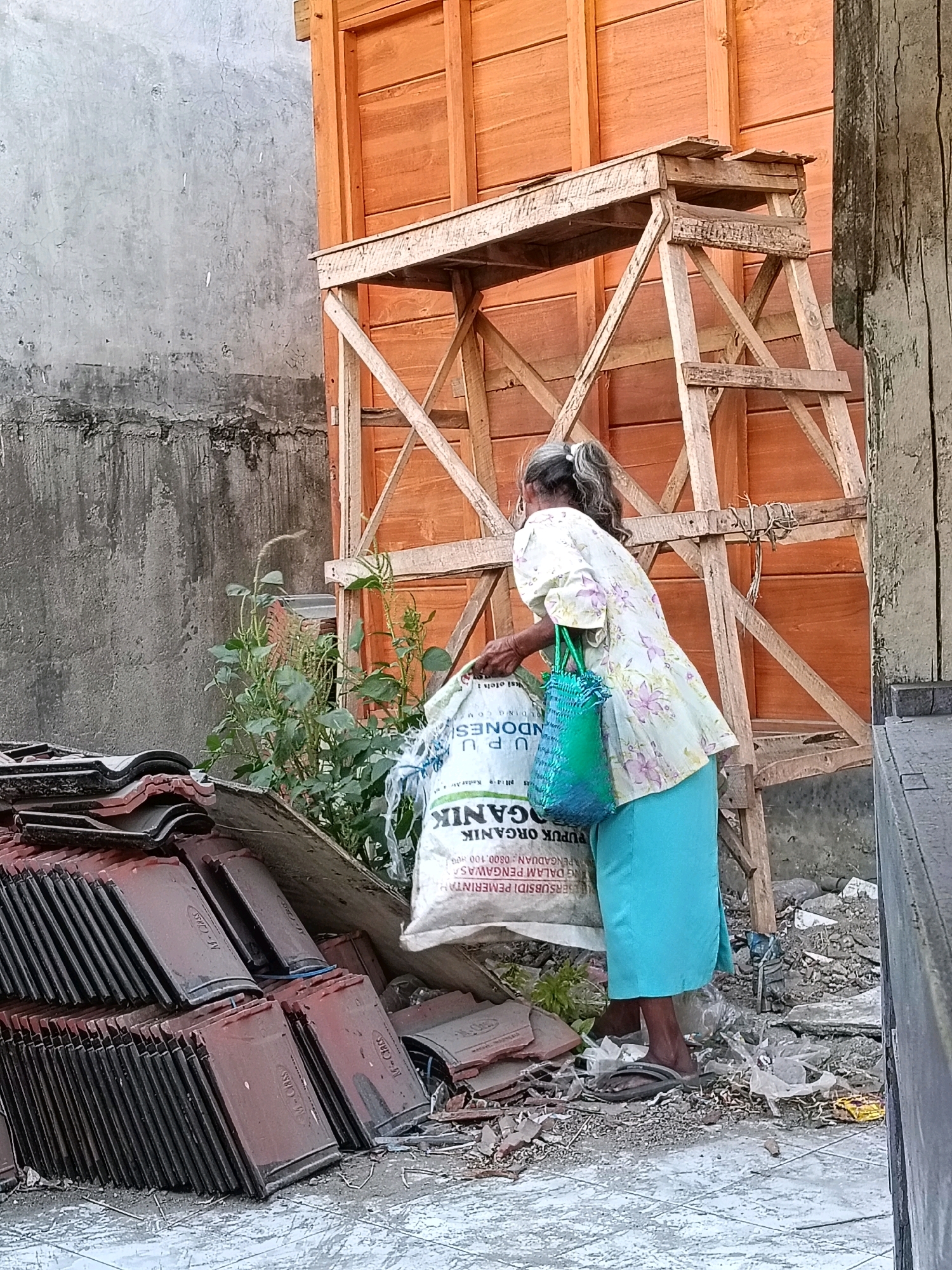 In Kedungtuban, Indonesia, a woman with graying hair tied in a ponytail stands outdoors, facing away from the camera. She wears a light-colored blouse with a floral pattern and a bright blue skirt. She is holding a large white bag with text in Indonesian, which appears to be fertilizer, and a smaller green woven bag. The scene is cluttered with construction debris, including stacks of red roof tiles and discarded materials. In the background, a wooden structure, possibly scaffolding or part of a building under construction, is visible, along with a textured gray wall. The ground is covered in a mix of dirt, small stones, and scattered debris, with a hint of a wet, reflective surface on the lower right. The lighting suggests it is daytime, possibly late afternoon due to the warm tones of the wooden structure.