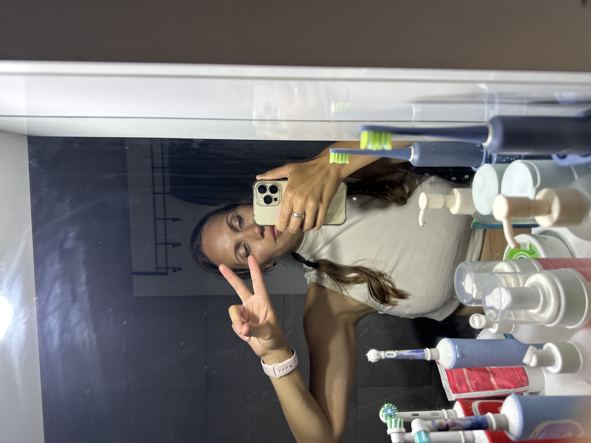 A woman takes a selfie in a bathroom mirror. She is holding her phone with her right hand, displaying a peace sign with her left hand, and wearing a pink smartwatch. Behind her, a shelf holds several electric toothbrushes, toothpaste tubes, and bottles of soap or lotion. The bathroom has gray tiles and a white countertop. While the specific location in Fethiye, Turkey, is not discernible from the image, it can be inferred that the photo was taken in a private residence or hotel bathroom in Fethiye.