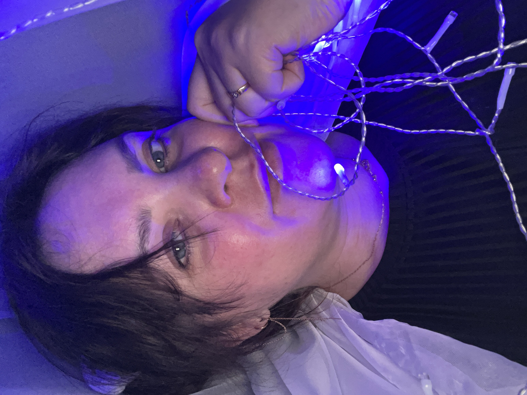 A medium close-up shot features a fair-skinned individual with dark hair, likely female, illuminated by blue LED fairy lights. The person is reclined, with their face angled towards the camera. They are holding a string of these lights near their face, specifically their cheek. A gold ring is visible on their hand. The subject's eyes appear to be light-colored. They are wearing a white garment and a gold necklace. The background appears dark. The location in the city of Noviy, Russia is unknown.