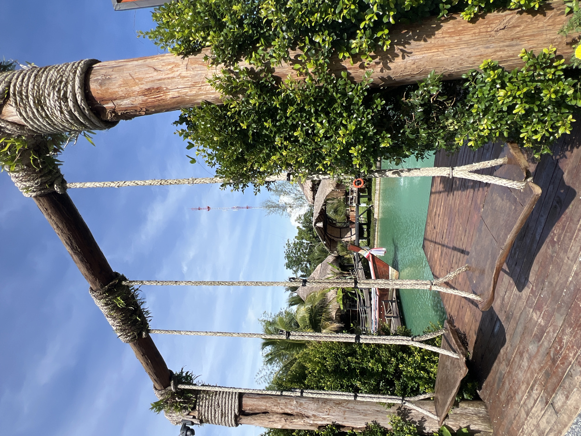 This image depicts a rustic outdoor scene with two wooden swings suspended from a sturdy wooden beam, supported by thick wooden posts wrapped in rope. The swings hang over a wooden deck, which leads to a tranquil body of turquoise water. In the background, traditional thatched-roof huts and palm trees suggest a tropical setting. A longtail boat is partially visible on the water, adding to the local ambiance. The scene is set against a bright blue sky with scattered white clouds. The overall impression is one of a peaceful, resort-like environment, likely in a location like Tambon Pa Kae Bo Hin, Thailand, known for its scenic beauty and relaxed atmosphere.