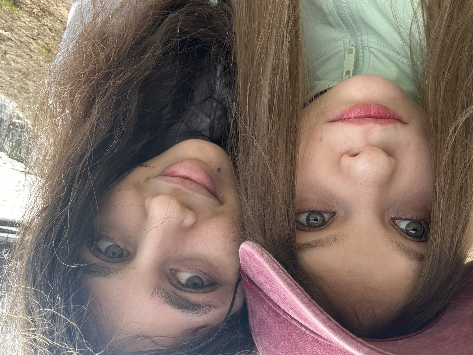 Two young women are closely positioned, faces near each other. The woman on the left has dark brown hair and is wearing a dark colored garment. The woman on the right has light brown hair and is wearing a light green jacket. Part of a pink hat is visible near her face. The background is blurry and indistinct, appearing to be an outdoor environment. The image is rotated so that the faces are upside down. There is no contextual information to suggest this image was taken in Moscow, Russia.