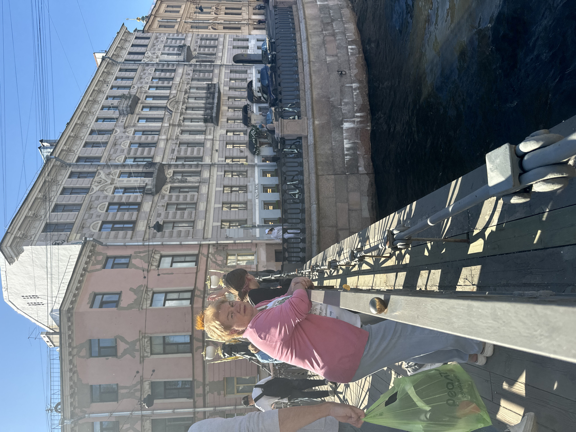 In Saint Petersburg, Russia, an elderly woman with blonde hair, wearing a pink shirt and grey pants, stands on a bridge overlooking a canal. She is holding a bright green plastic bag. Behind her, other people are visible, some leaning on the bridge railing. The scene is bathed in sunlight, suggesting a clear day, likely daytime. The background features grand, old buildings with ornate facades lining the canal. The architecture suggests a historic European city. The water in the canal is dark and appears to be flowing. A prominent feature of the bridge is its sturdy metal railing with many vertical supports. The overall impression is of a pleasant day out in a picturesque urban environment.