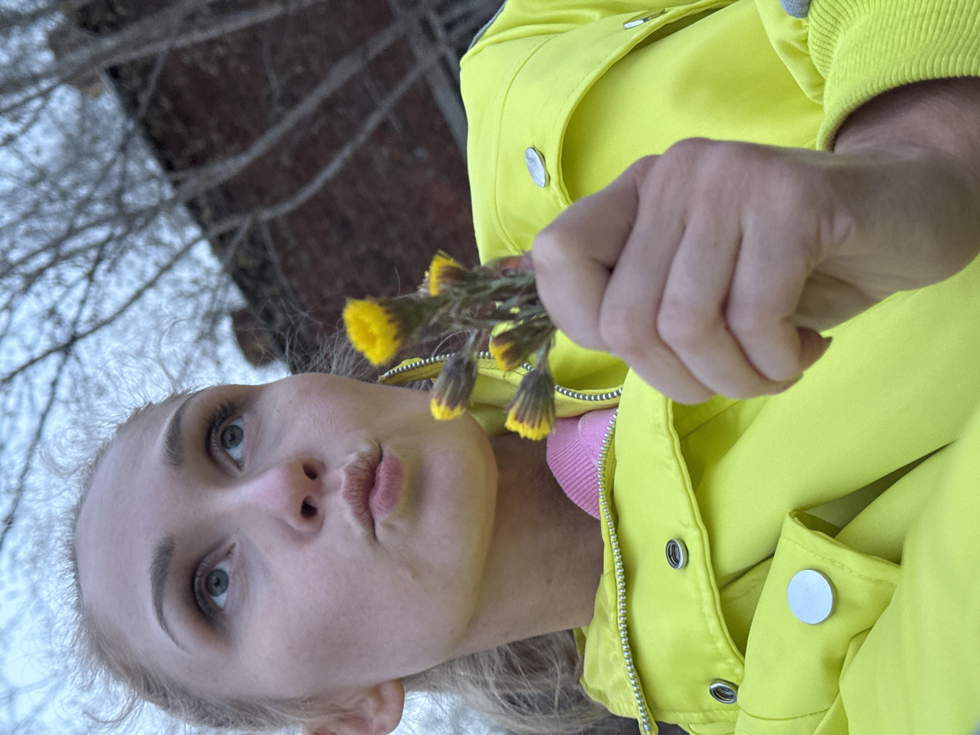 A young woman with blonde hair and light-colored eyes is captured in an outdoor, close-up shot, angled slightly upwards towards the camera. She holds a small bunch of yellow flowers, resembling coltsfoot, in her right hand, positioned near her face. Her lips are slightly pursed, and her gaze is directed forward.
She is wearing a bright, almost neon, yellow jacket with silver snap buttons and a prominent silver zipper. A pink garment is visible underneath the jacket. Her fair skin shows minimal makeup, highlighting her eyebrows and eye makeup.
The background is blurred, featuring bare tree branches against a light, overcast sky or snowy ground, suggesting an early spring or late winter setting. A dark, textured surface, possibly a brick wall or building, is also visible in the upper background. The presence of coltsfoot flowers, which bloom early in the season, aligns with this temporal context. The overall mood of the image is calm and natural. No visible text is present. Given the location context, this scene is in Chaikovsky, Russia.