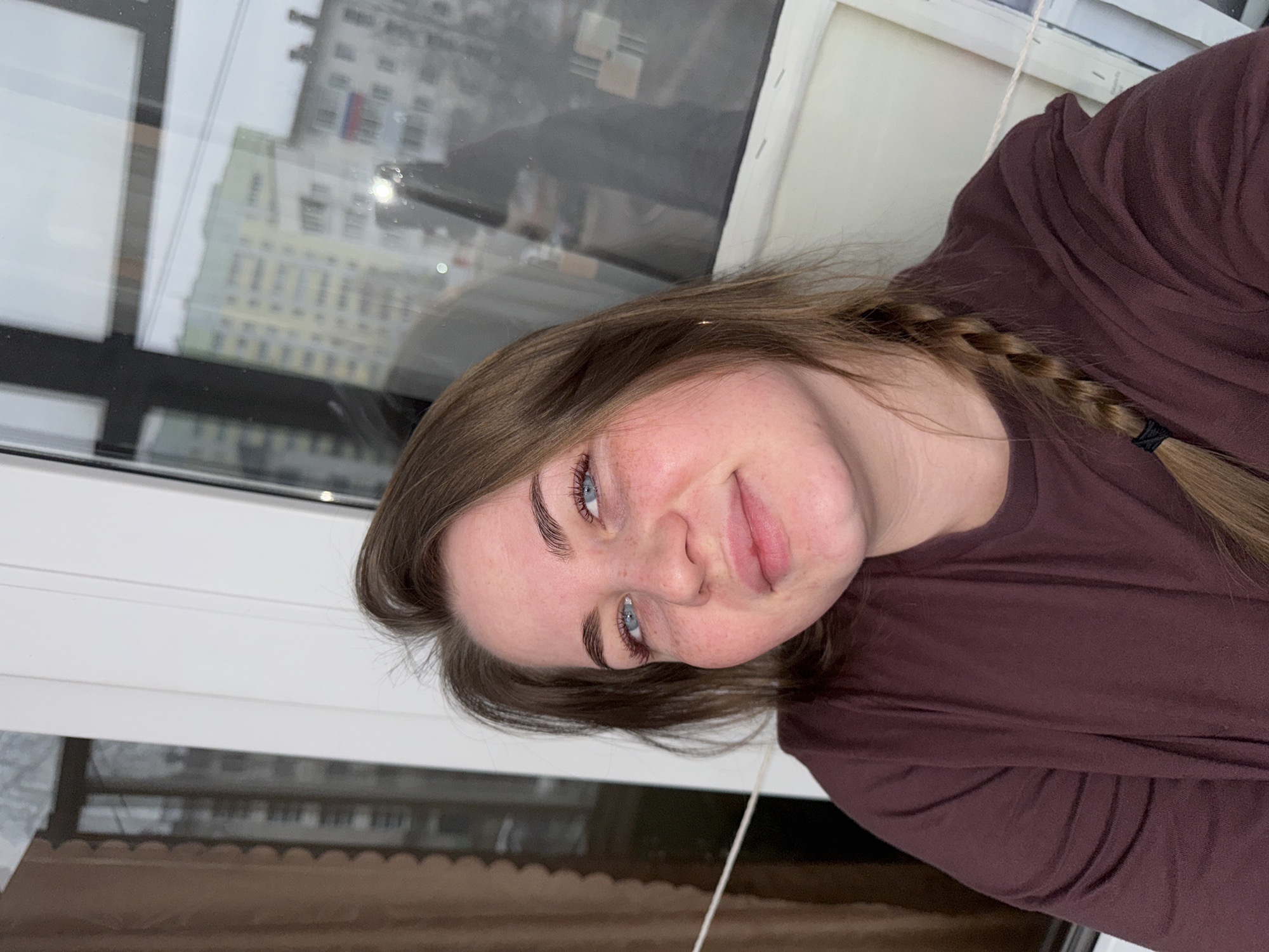 A woman with blue eyes and brown hair is looking at the camera with a slight smile. Her hair is styled with a braid on one side. She is wearing a dark purple shirt. The background shows an out-of-focus urban landscape visible through a window. It is possible to identify a building in the city of Chaikovsky, Russia, based on the typical architecture of the buildings. The setting appears to be indoors, near a window.
