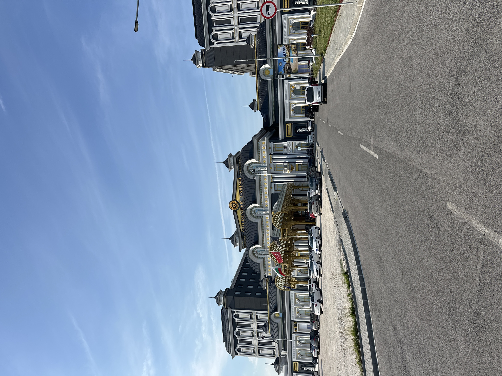 This image captures the exterior of the "GOLDENEYE CASINO" complex in Kapitan Andreevo, Bulgaria, under a clear blue sky with sparse, wispy clouds. The casino building is a large, ornate structure featuring a neoclassical architectural style with dark gray/black facades accented with gold trim and white columns. Multiple dome-like structures, some topped with spires, are visible across the roofline. The central section of the building prominently displays the "GOLDENEYE CASINO" logo in gold above a grand entrance with a golden arched canopy. Several classical statues in white are positioned near the entrance and along the building's facade.
In front of the main entrance, two flags are displayed: the national flag of Bulgaria and the national flag of Turkey. A banner on the right side of the complex advertises "YAZ AYRICALIĞI" (Summer Privilege) with "25€" and an image of a pool.
The foreground features a paved asphalt road leading directly towards the casino entrance, marked with white dashed lines. A white delivery van is visible on this road, facing the building. To the left of the road is an unpaved gravel parking area occupied by multiple parked cars of various makes and colors, predominantly white, silver, and dark. To the right of the road, a sidewalk and a strip of manicured grass with low vegetation are present. A lamppost stands on the right, and a red circular road sign with a white truck symbol and a diagonal line, indicating "no trucks" or "trucks prohibited," is clearly visible. Another section of the building, similar in architectural style, extends to the far right. The overall scene appears well-maintained and devoid of any prominent human figures.