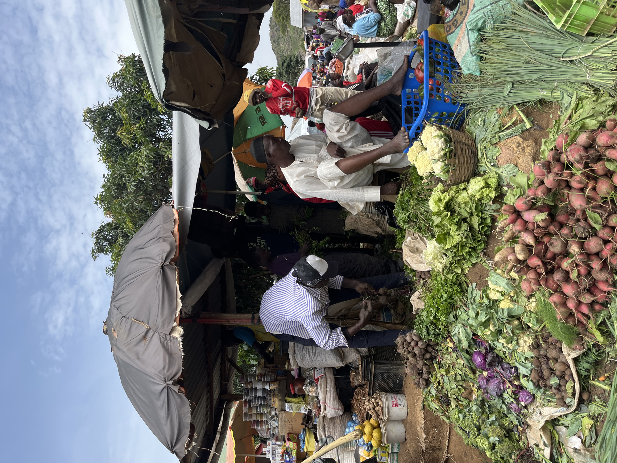 This image depicts a bustling outdoor market scene in Gura Top, Nigeria. In the foreground, a man wearing a traditional white robe and skullcap sits on the ground, facing to the left, appearing to be engaged in conversation or observing the market. To his right, a pile of fresh vegetables, including radishes, cauliflower, leafy greens, and what appear to be leeks, is displayed. Behind him, another man in a red hoodie stands, holding some produce. Further in the background, the market extends with more stalls, people, and a variety of goods. The setting is open-air, with makeshift shelters and umbrellas providing shade. The sky is visible in the upper left corner, suggesting it is daytime. The overall atmosphere appears lively and busy, characteristic of a local marketplace. There is no visible text in the image.