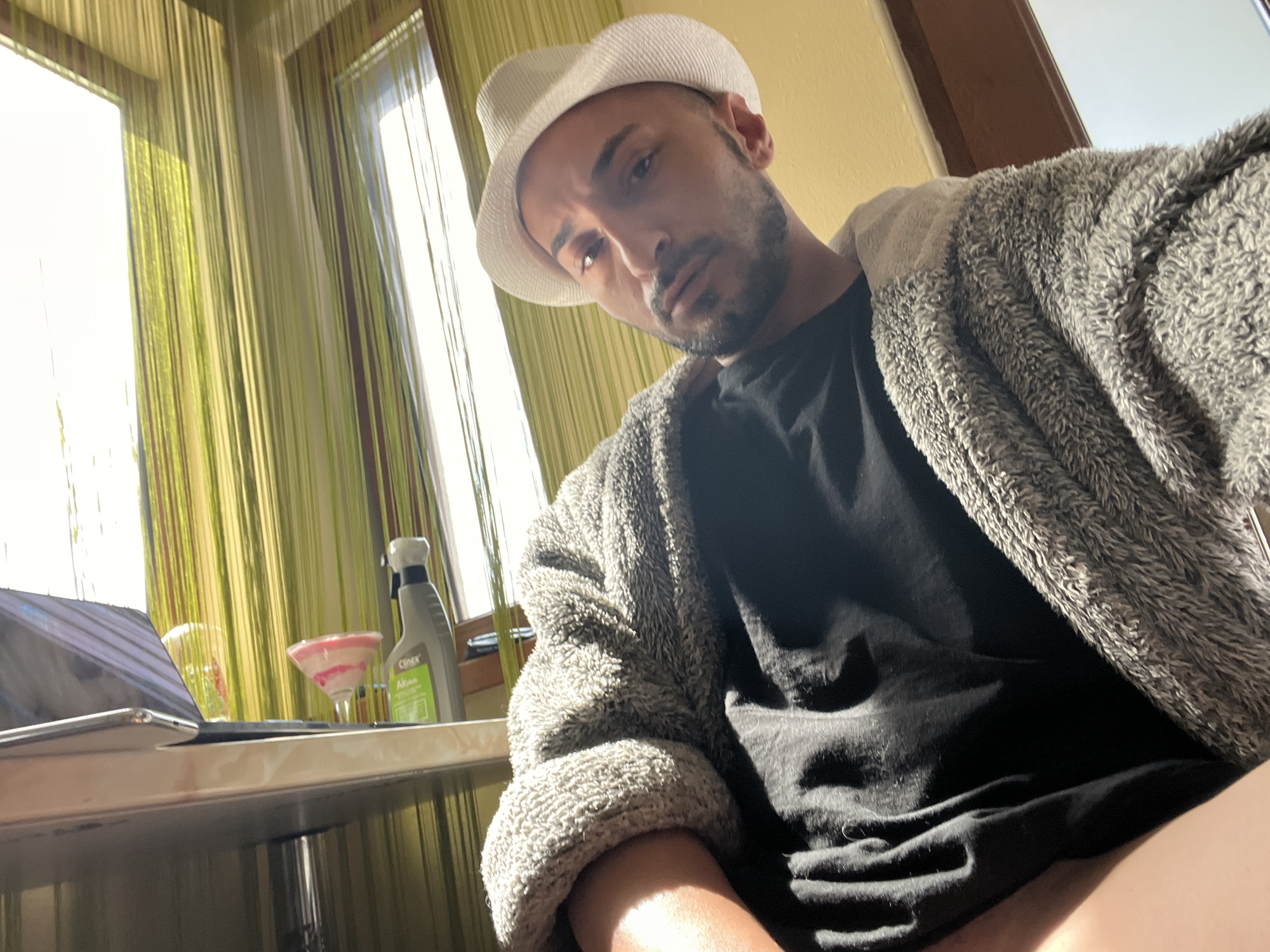 A man in his late 20s or 30s poses for a selfie indoors in Medkovets, Bulgaria. He wears a white fedora-style hat, a black t-shirt, and a grey, textured (possibly fleece or chenille) robe or cardigan, looking directly at the camera with a serious or pensive expression. He has short facial hair.
Bright natural daylight streams through two large windows in the background, which are covered with distinctive vertical string curtains in shades of green and yellow. The room's walls appear to be a light yellow or beige. On a surface, possibly a table or shelf, to the viewer's left, a silver laptop or tablet is partially visible. Next to it are a clear glass with a red rim and a cocktail glass holding a pink, swirled beverage. A grey spray bottle with a white nozzle, labeled "Clinex All-plus," is also present on the surface. The image is taken from a low angle, giving a casual, relaxed vibe during the daytime.