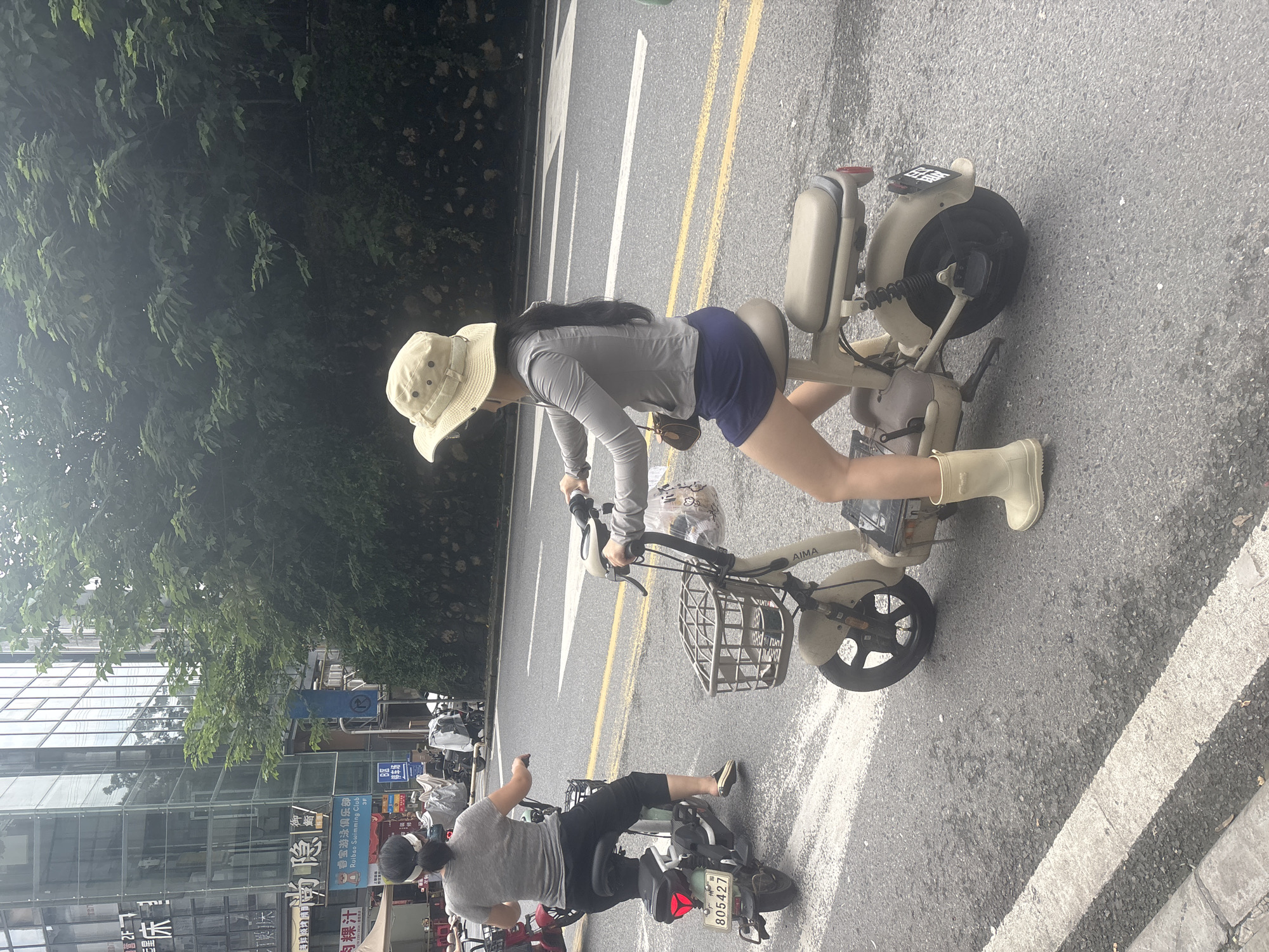 In Nanhai District, China, two individuals are operating electric scooters on a street. In the foreground, a person wearing a wide-brimmed hat, a grey long-sleeved shirt, and a dark blue skirt rides a beige electric scooter. They are also wearing light-colored boots and have a small brown purse slung over their shoulder. In the mid-ground, a second person, facing away from the camera, rides a red and white electric scooter. They are wearing a grey t-shirt and dark pants, with a white headband in their hair. The background shows a modern building with glass windows and a lush green tree-lined wall. The street is paved and marked with white and yellow lines, indicating a road.