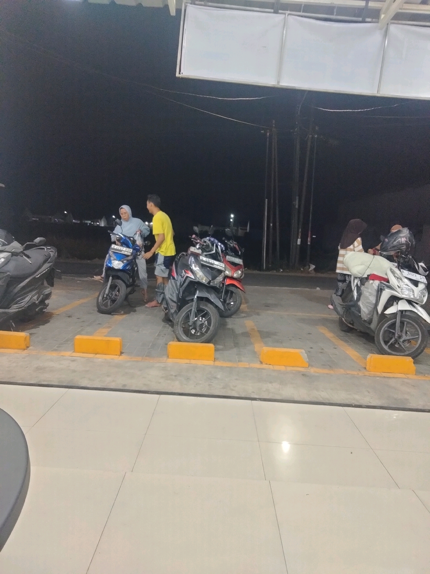 At night, in a parking area in Suka Sari, Indonesia, a man in a yellow t-shirt and shorts converses with a woman wearing a blue hijab and light-colored pants. They stand next to a blue scooter. Several other scooters are parked nearby, including a black one on the left, a black and red one, and a white one on the right. The parking spots are marked with yellow bollards. The background consists of utility poles and a dark sky, with some overhead lighting structures. The ground is paved with concrete and tiles.