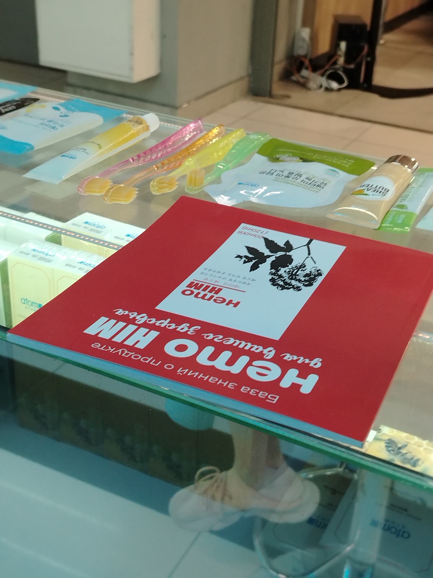 The image depicts a glass display case containing various cosmetic and personal care products. A red book with white Cyrillic text and an image of a plant is prominently placed in the foreground. The text on the book includes "HEMO HIM". Behind the book are several toothbrushes with yellow and pink handles, along with tubes and packets of cosmetic products bearing labels with the word "atom" or "atomy" in English and Korean.
The display case appears to be in a shop or store setting. The location is inferred to be Izhevsk, Russia, based on the Cyrillic text.