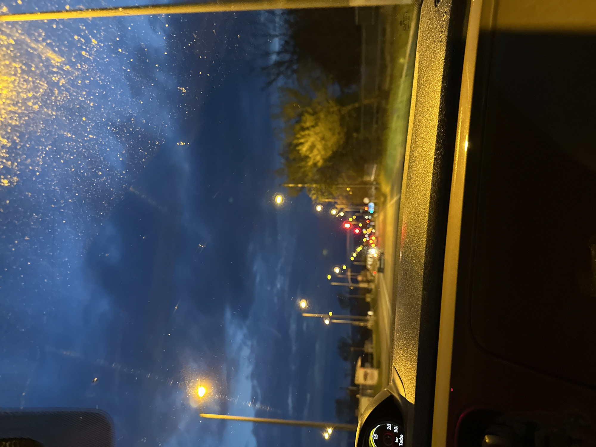 The image depicts a nighttime scene viewed through a car windshield in Lefkonas, Greece. The windshield is somewhat dirty with streaks and spots. A street lined with evenly spaced streetlights stretches into the distance, leading towards a cluster of red and white lights, likely indicating a traffic intersection. Vegetation can be seen along the roadside. The car's dashboard is visible in the lower portion of the frame, with a portion of the instrument cluster showing gauges and indicators. The overall environment suggests a slightly overcast or cloudy night.