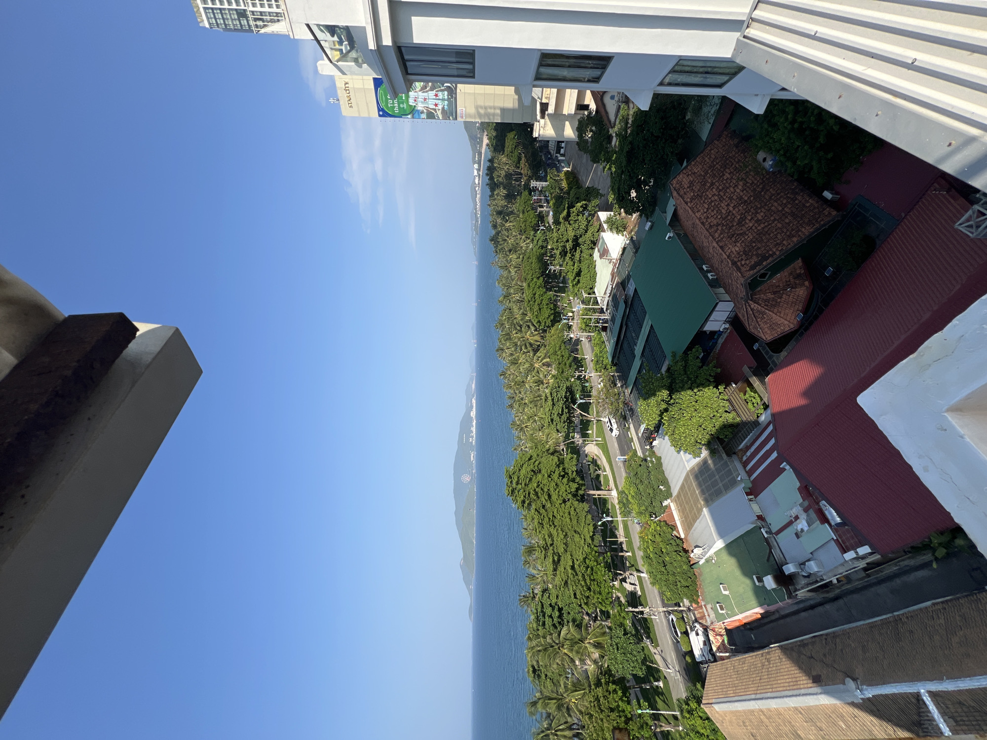 The image presents a high-angle view of a coastal city, likely Nha Trang, Vietnam. The foreground is dominated by the white railing of a balcony and a portion of a building. Below, a densely packed urban landscape unfolds, characterized by various buildings with diverse roof types, including red tile, green metal, and some covered with tarpaulin. A road runs through the city, with a few cars visible, including a white sedan and a white van. Lush green trees, including palm trees, line the road and are interspersed throughout the urban area.
Beyond the city, a wide expanse of the blue sea stretches towards the horizon. In the distance, a large, green, hilly island is visible, with some structures and what appears to be a Ferris wheel on its peak. The sky is a clear, vibrant blue, with a few wispy white clouds. A tall, white building with multiple floors and visible windows is prominent on the right side of the frame, displaying a large advertisement. Another taller building with "STAR CITY" signage is also visible further down the coast. The overall scene depicts a sunny day in a tropical coastal city.
