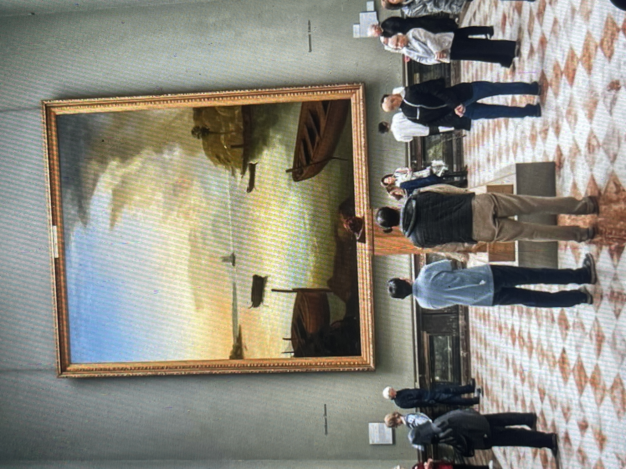 The image depicts a group of people viewing a large, framed painting of a seascape with boats and a coastline, hung on a pale grey wall in a museum. The painting features a dramatic sky with clouds at sunset or sunrise, casting warm light on the water and land. Several boats are visible in the water, some moored and others in motion. The floor is tiled in a diamond pattern of white and reddish-brown squares.
In the foreground, three individuals are the primary subjects, all facing away from the camera towards the painting. The person on the left is wearing a dark hooded jacket and dark trousers. The person in the center is wearing a light blue collared shirt and dark trousers. The person on the right is wearing a dark vest over a light-colored shirt and light brown trousers.
Behind them, other museum visitors are scattered, some also observing the artwork. These include individuals in the middle ground and background, some standing, others appearing to converse. The overall impression is of a public exhibition space. The image does not provide any visual cues or context to suggest the location is Bishkek City, Kyrgyzstan.