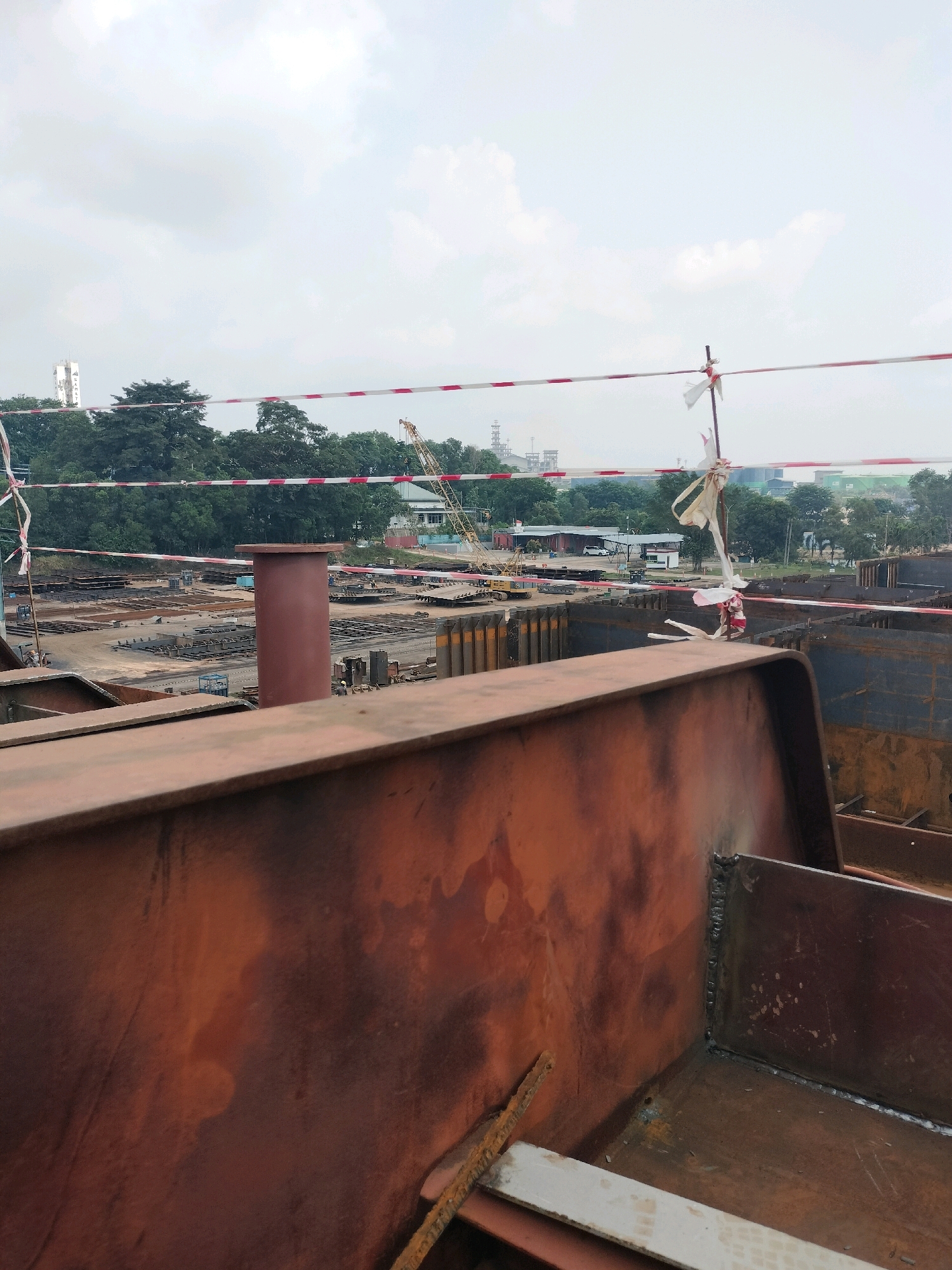 The image captures a construction site, likely a shipyard or heavy industrial facility, in Batam, Indonesia. In the foreground, large, weathered steel structures, possibly ship sections or industrial components, dominate the frame. These are rusted and show signs of welding. Behind these, a wide expanse of the construction yard is visible, filled with stacks of steel plates, beams, and pre-fabricated sections laid out on the ground.
Several cranes are present in the mid-ground, including a prominent yellow crane with its boom extended. Smaller vehicles, including what appears to be a yellow mini-excavator, are also visible. A cylindrical, dark red structure, resembling a large pipe or pillar, stands in the mid-left of the image.
A red and white caution tape is strung across the scene, supported by poles, indicating a restricted area. In the background, lush green trees form a treeline, separating the industrial site from the distance. Beyond the trees, industrial buildings with smokestacks are visible, suggesting proximity to other manufacturing or processing facilities. The sky is overcast with light clouds. The overall scene is one of ongoing industrial construction and activity.