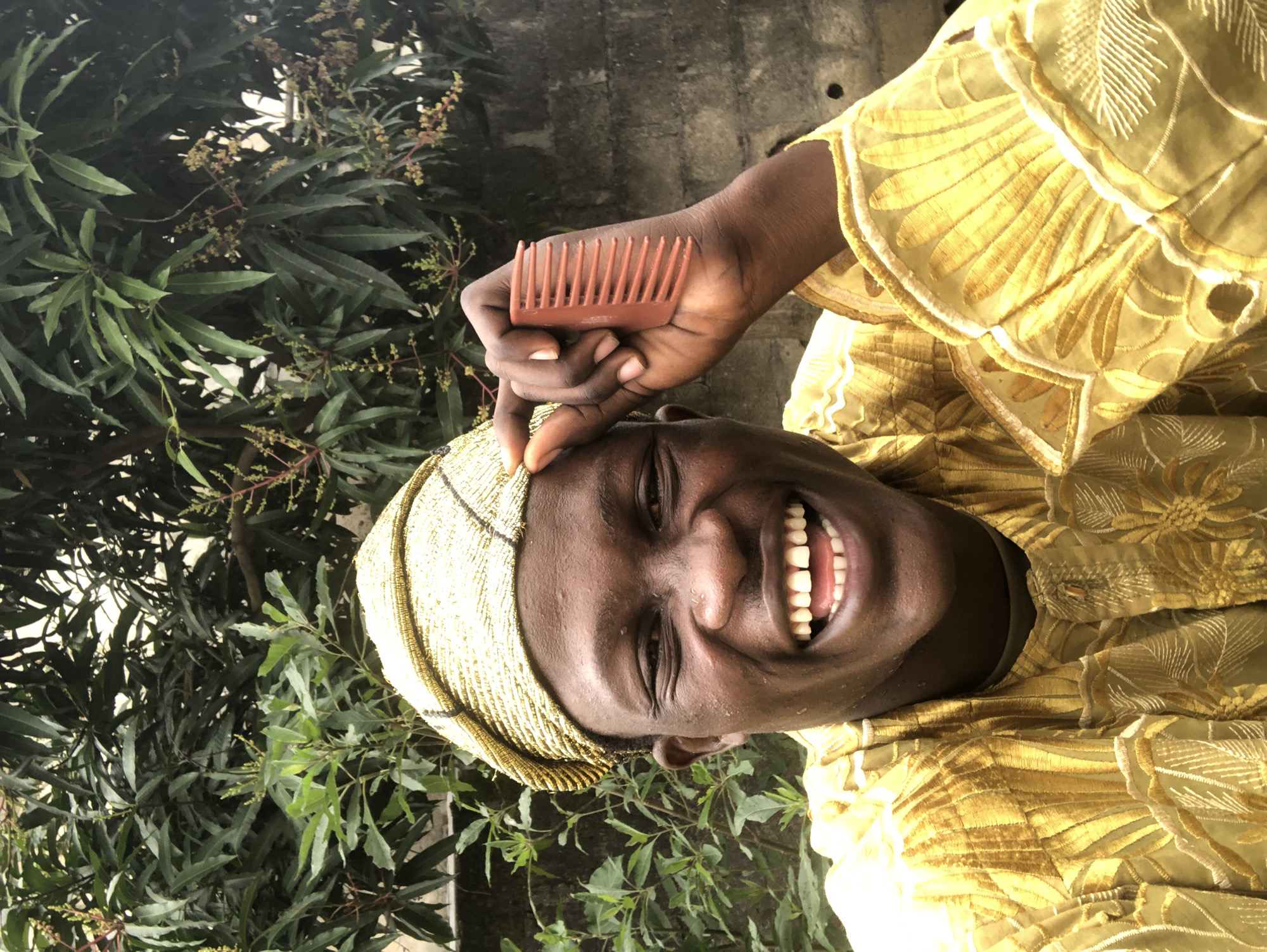 A close-up, waist-up shot shows a Black man in Ado, Nigeria, smiling broadly while wearing a traditional golden embroidered outfit and a matching cap. He holds a small, orange comb to his temple with his right hand. The background features lush green foliage and a textured grey wall, suggesting an outdoor setting in a residential area. The man's expression is joyful and animated, with his eyes crinkled at the corners. His teeth are visible, and his mouth is open in a wide laugh. The intricate details of his clothing, with its elaborate stitching and metallic sheen, are prominently displayed. The natural lighting creates highlights and shadows on his face and attire, adding depth to the image. The overall impression is one of warmth, happiness, and cultural pride.