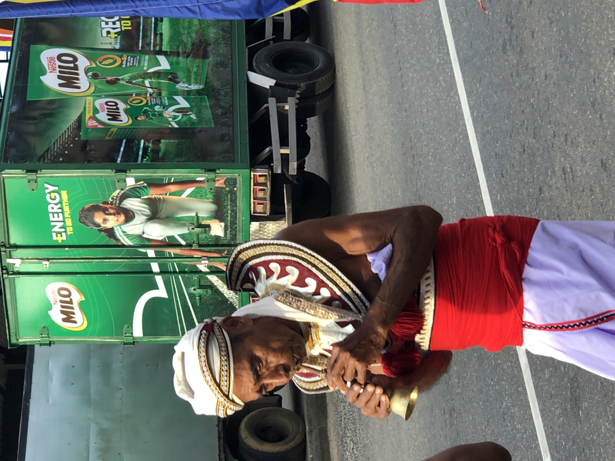 A man in traditional Sri Lankan attire, including a white turban with gold and red trim and a red sash, plays a small brass instrument. He is standing in front of a green truck adorned with advertisements for Nestle Milo. The truck appears to be a mobile promotional vehicle, featuring images of athletes and the Milo logo. The scene takes place outdoors on a paved surface, possibly a road, with a white line marking a lane. The man's expression is focused on his playing. The overall atmosphere suggests a public event or promotional activity. The visible text on the truck includes "Nestle MILO" and "ENERGY TO GO FURTHER." The location context provided is Habaraduwa, Sri Lanka, implying this is a local event. The lighting suggests it is daytime.