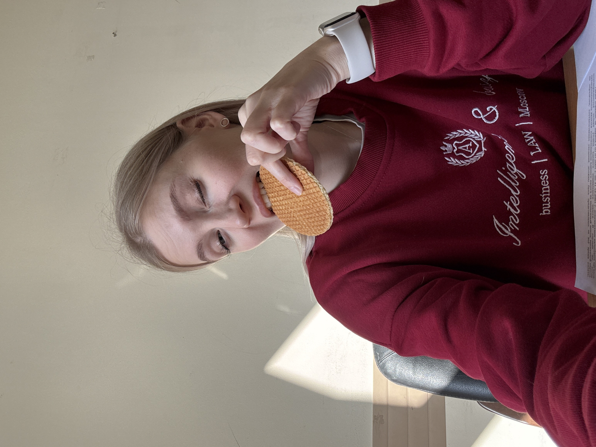 A woman is holding a stroopwafel in her right hand, and putting it near her mouth. She is wearing a maroon sweatshirt that reads "Intelligen business | LAW Moscow", and she has an Apple Watch on her left wrist. The scene appears to be indoors with a cream-colored wall visible in the background. The location is not possible to determine as Izhevsk is not mentioned in the OCR of the image.