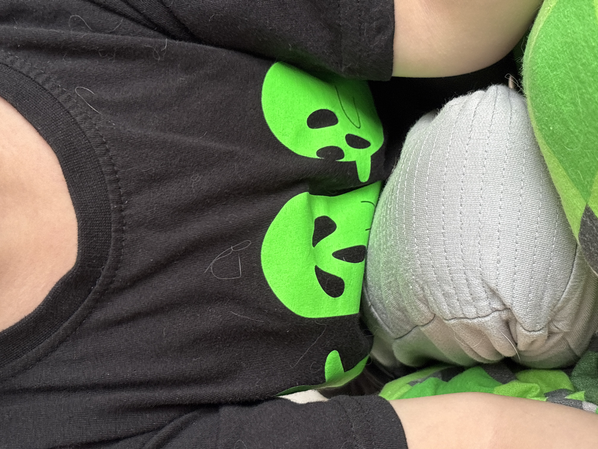 A person wearing a black t-shirt with a repeating green alien head design is lying down. They are holding a stuffed grey object, possibly a pillow or toy, against their body. A green blanket is partially visible next to the stuffed object. The scene does not contain enough information to locate it in the city of Ембаево, Russia.