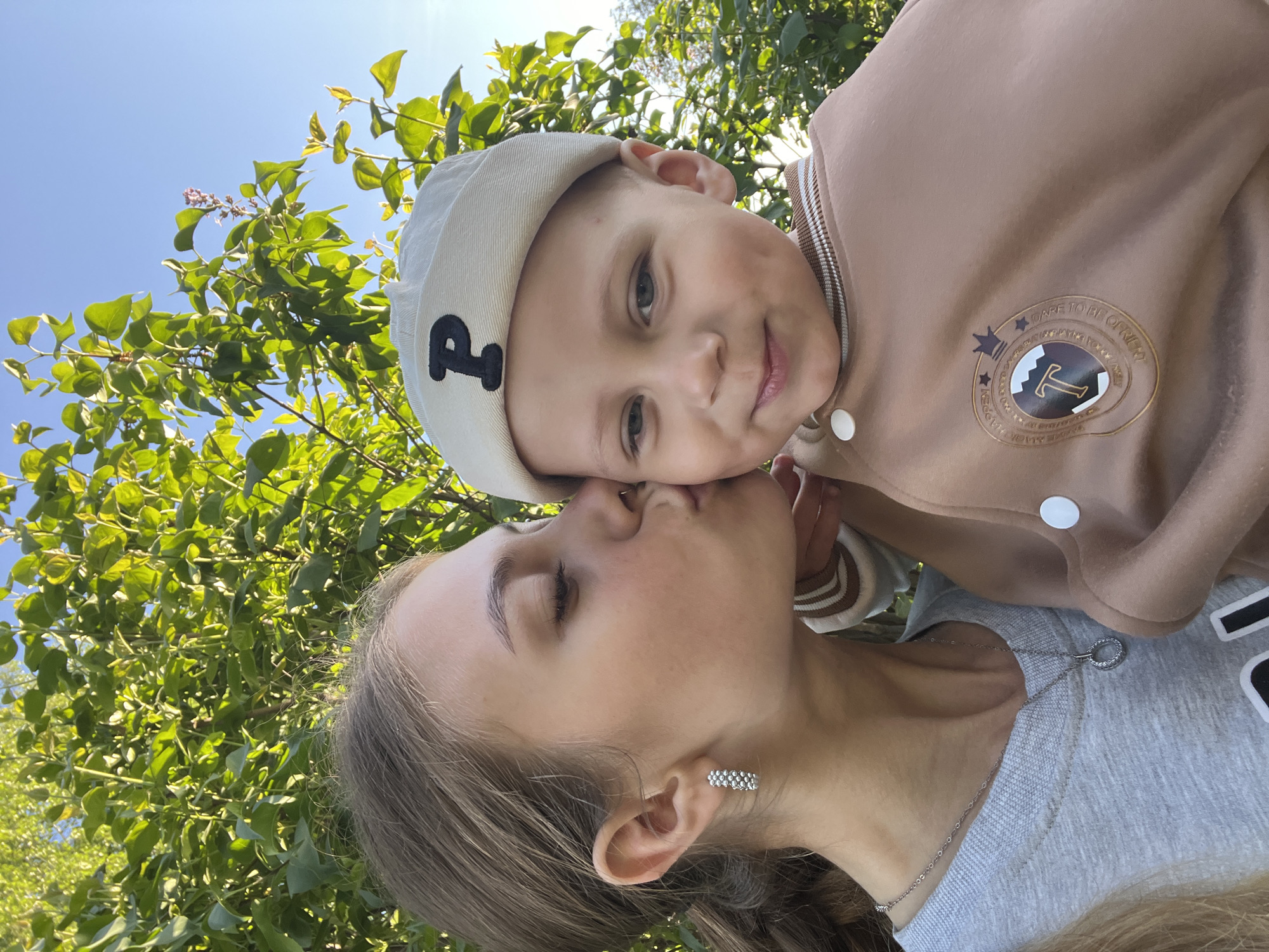 A woman with light brown hair and silver earrings kisses a young boy on the nose. The boy is wearing a beige jacket with a "T" emblem and a white cap with a "P" on it. They are outdoors, surrounded by green trees and a blue sky. It is not possible to confirm the location as Фоки, Russia from the given image.