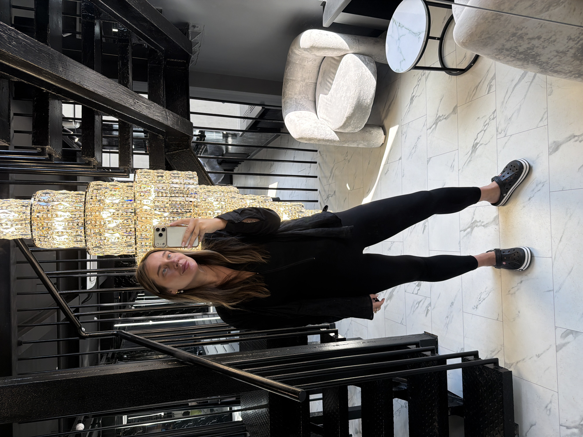 In a dimly lit, modern interior with marble-patterned flooring and a striking multi-tiered crystal chandelier, a woman with long brown hair stands facing a mirror, taking a selfie with her smartphone. She is dressed in a black V-neck zippered top and black leggings, accessorized with black Crocs. To her left, a sleek black metal staircase with open risers ascends, its railing mirroring the vertical lines of the chandelier. To her right, a plush, light gray armchair with a curved seat and thick legs sits near a small, round side table with a white marble top and black metal base. The background reveals more of the staircase structure and dark-paneled walls, suggesting a chic, possibly residential or commercial space. The lighting, originating from the chandelier and possibly other unseen sources, casts dramatic shadows and highlights on the scene.